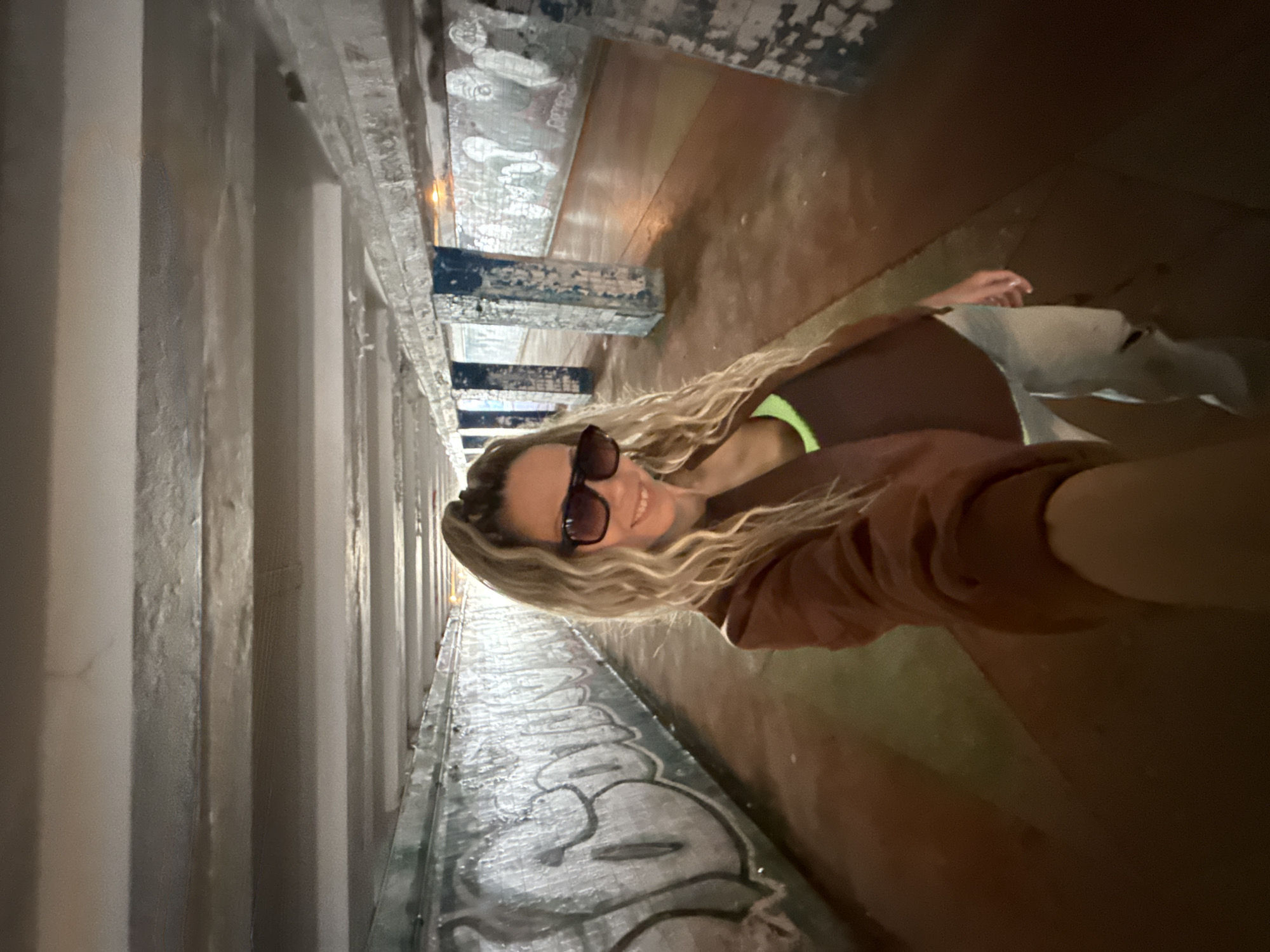 A blonde woman with long, wavy hair and sunglasses takes a selfie in a concrete underpass. She is wearing a brown jacket over a neon green and brown top and ripped light-wash jeans. The underpass walls are covered in graffiti. The floor is a reddish-brown color. The scene is likely in Zhukovo, Russia.
