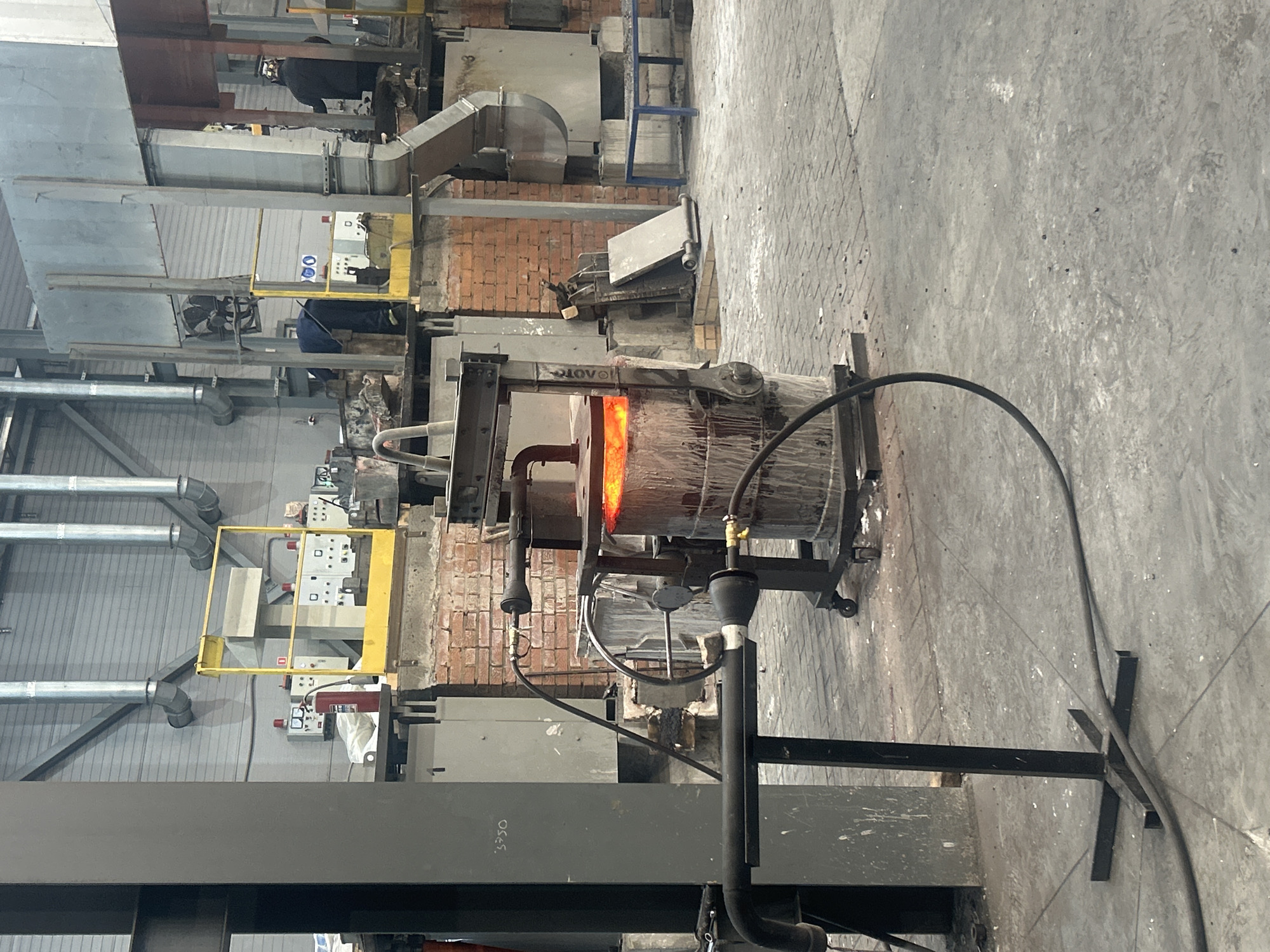 The image depicts an industrial setting in Noviy, Russia, likely a factory or manufacturing plant. The focus is on a furnace, with a section of it glowing intensely orange, indicating high heat. The furnace is supported by a complex metal framework. Various pipes and ducts run throughout the scene, typical of industrial infrastructure. Brickwork is visible around the furnace area, suggesting insulation or containment. A cable extends from the furnace towards the foreground, attached to a support structure with a stand on the concrete floor. In the background, there are glimpses of control panels, machinery, and metal structures. The overall impression is of a functional, but possibly dated, industrial workspace.