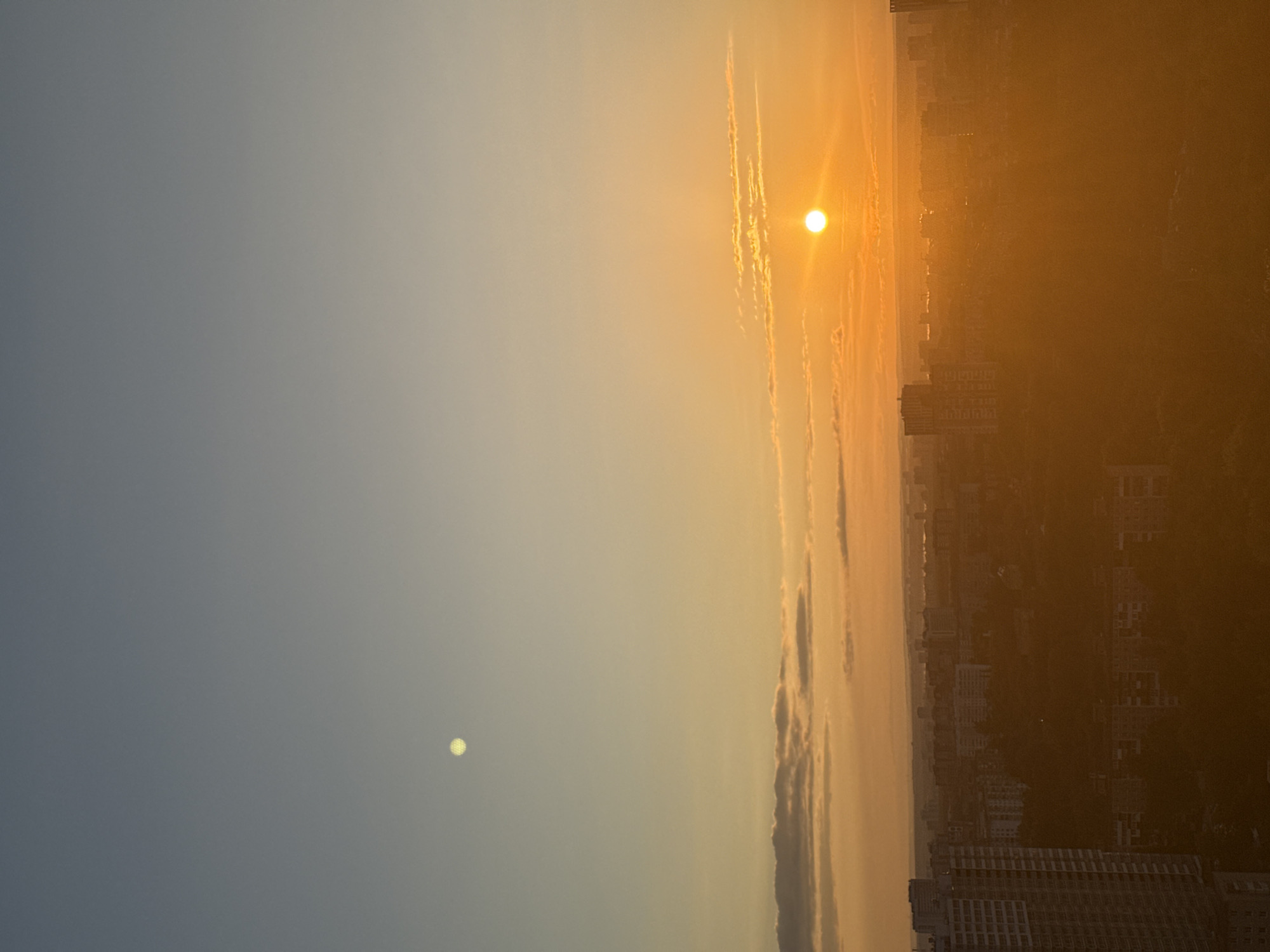 The image presents a panoramic view of either a sunrise or sunset over an urban landscape, likely Perm, Russia. The sky dominates the frame, displaying a striking gradient of colors from a deep, cool blue-grey on the left to a vibrant, warm golden-orange on the right.
A brilliant, intense sun is positioned in the upper right portion of the frame (if rotated to landscape orientation), casting a powerful, radiant glow that illuminates the clouds and the distant city. Thin, wispy clouds, elongated and streaked with golden light, stretch horizontally across the middle of the sky, catching the sun's rays and adding texture to the expansive vista.
In stark contrast to the intense sun, a smaller, dimmer, yet distinct bright yellow-white spot is visible in the upper left section of the cooler, blue-grey sky. This could be the moon or a very bright star/planet.
Below the dramatic sky, the silhouette of a sprawling cityscape extends across the lower part of the image. Numerous multi-story buildings are visible, appearing hazy and somewhat indistinct due to the atmospheric conditions and the backlight from the low sun. The city is bathed in a warm, golden hue, creating a soft, almost ethereal ambiance as it stretches towards the distant horizon, which is also shrouded in a warm, golden haze.
The scene captures the serene and majestic beauty of the golden hour, highlighting the vastness of the sky and the peaceful transition of day. There is no visible text in the image.