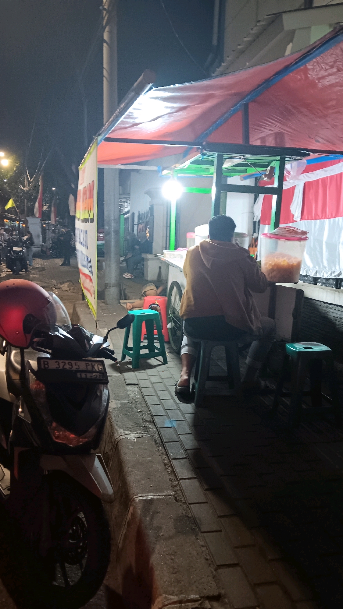 The image depicts an outdoor scene at night in South Jakarta, Indonesia, likely a street food stall. A person wearing a yellow hoodie and dark pants is seated on a green stool, facing away from the camera and towards a food preparation area. They are looking at a large clear container filled with yellow food items. Another person is seen lying on a red stool in the background, partially obscured. A red helmet is prominently displayed on the front of a parked motorcycle in the foreground, to the left of the frame. The street is paved with interlocking blocks, and a concrete curb separates it from the road. A red and white tarp covers the food stall, illuminated by artificial lighting, creating a warm glow. Indonesian flags are visible in the background, further confirming the location. The overall atmosphere suggests a casual, late-night setting.