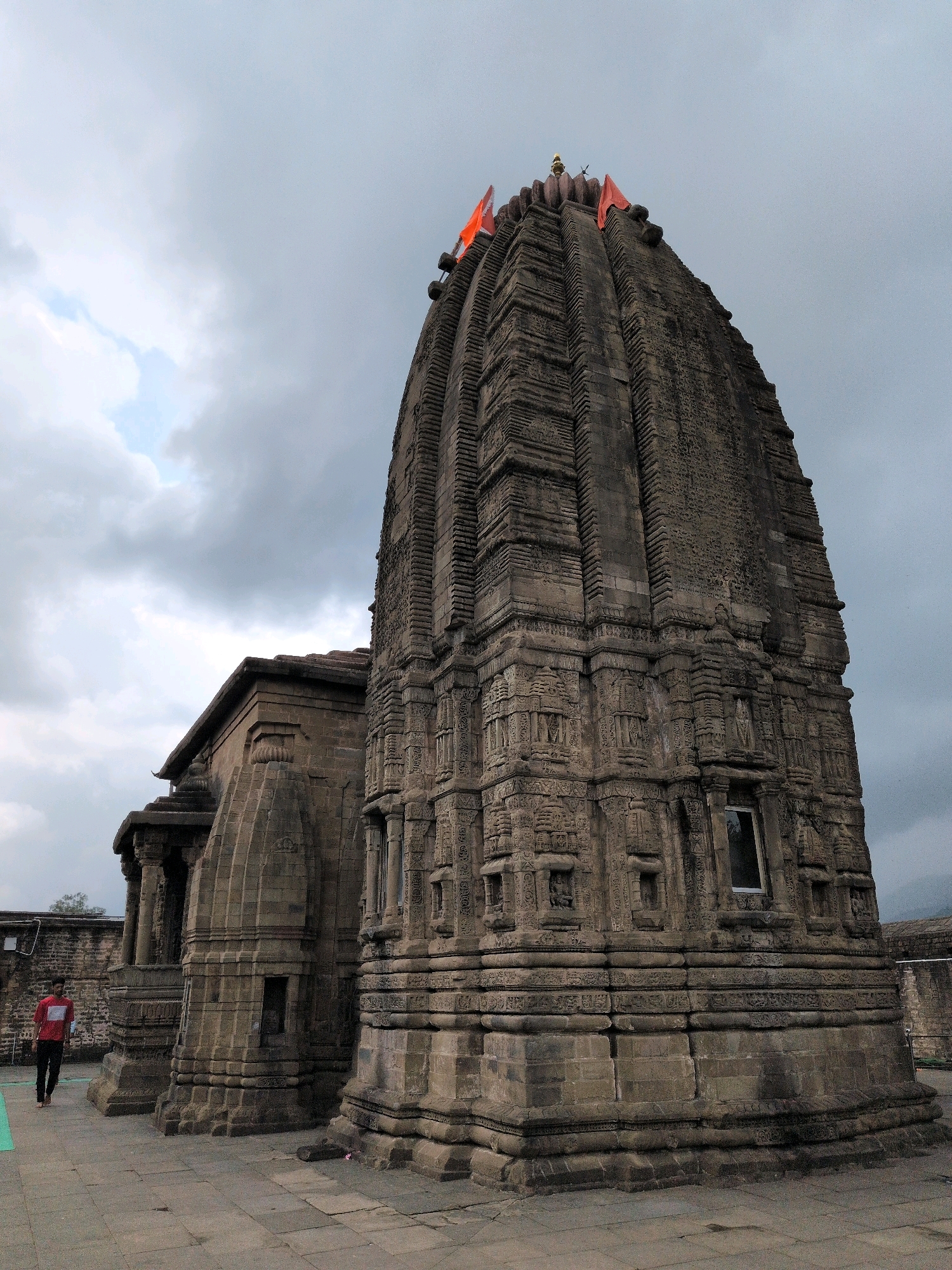 A side-view, low-angle shot captures a large, ancient stone temple in Baijnath, India, under a cloudy sky. The temple, with its intricately carved facade and tiered pyramidal roof, dominates the right side of the frame. Orange flags adorn the very top of the temple. To the left, a smaller, connected structure with a sloping roof is visible, also constructed from stone. In the foreground, a person wearing a red shirt and black pants walks away from the camera on a paved walkway. The ground is made of stone tiles. The background shows a stone wall and a hint of distant hills.