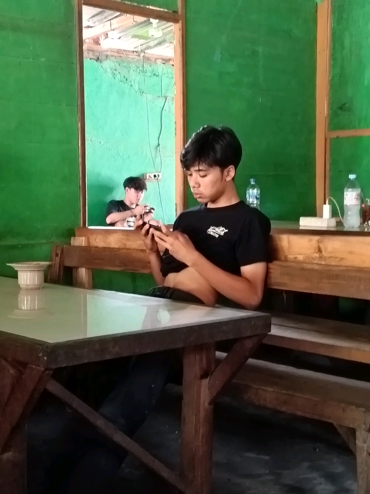 In Blora, Indonesia, a young man in a black t-shirt is seated at a wooden table, engrossed in his phone. His reflection is visible in a doorway behind him, where another young man is also looking at his phone. The room is painted green, and the furniture is made of wood. A bottle of water and a power strip are on a shelf behind the main subject. The overall scene depicts a casual, indoor setting.