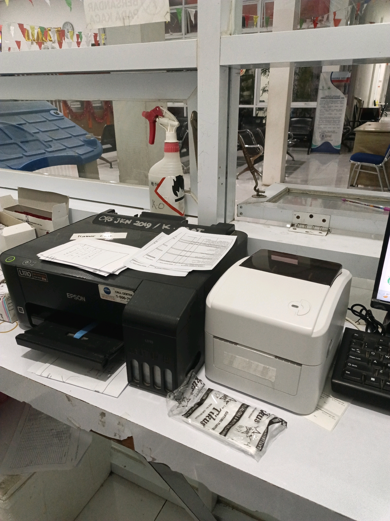 The image shows an office desk setting in Bangkalan, Indonesia. In the foreground, there is a black Epson printer, model L110, with several documents on top. To the right of the printer is a white thermal label printer. A black keyboard is partially visible to the right of the thermal printer. In front of the thermal printer, there are a few small, individually wrapped items with "TIRUS" branding. Behind the printers, a white spray bottle with a red nozzle is visible, with some labeling indicating "AL. CO" and "K.O". The desk is positioned near a window with a view of an interior space that appears to be an office or waiting area, with chairs and a banner visible in the reflection. The overall environment suggests a working office space.