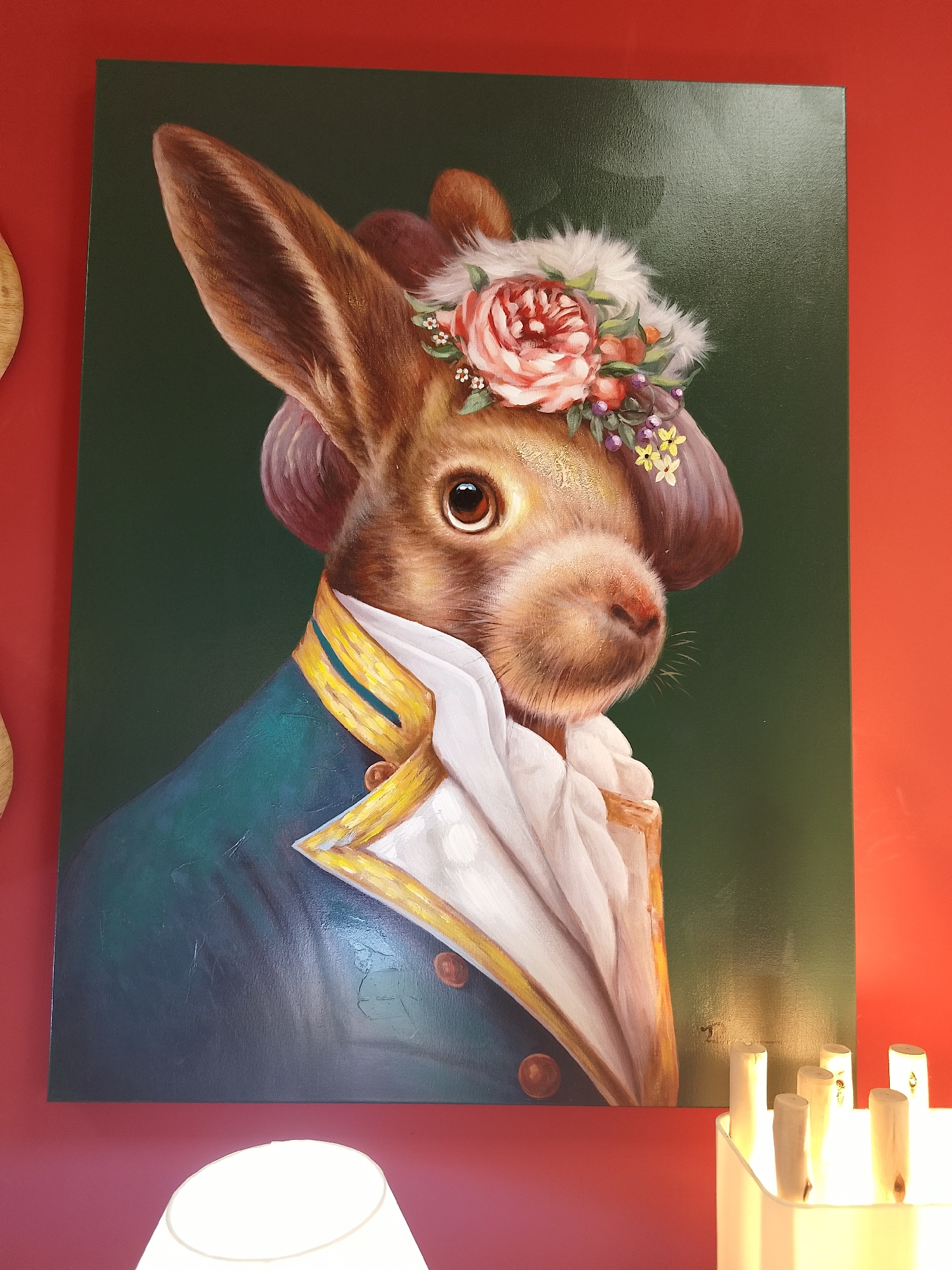 A painting of a rabbit dressed in formal attire hangs on a red wall. The rabbit wears a blue coat with gold trim and a white ruffled shirt. It also sports a floral headpiece. The painting is lit from below by a table lamp, which sits near a decorative container filled with wooden sticks. The location is Unknown, Unknown.