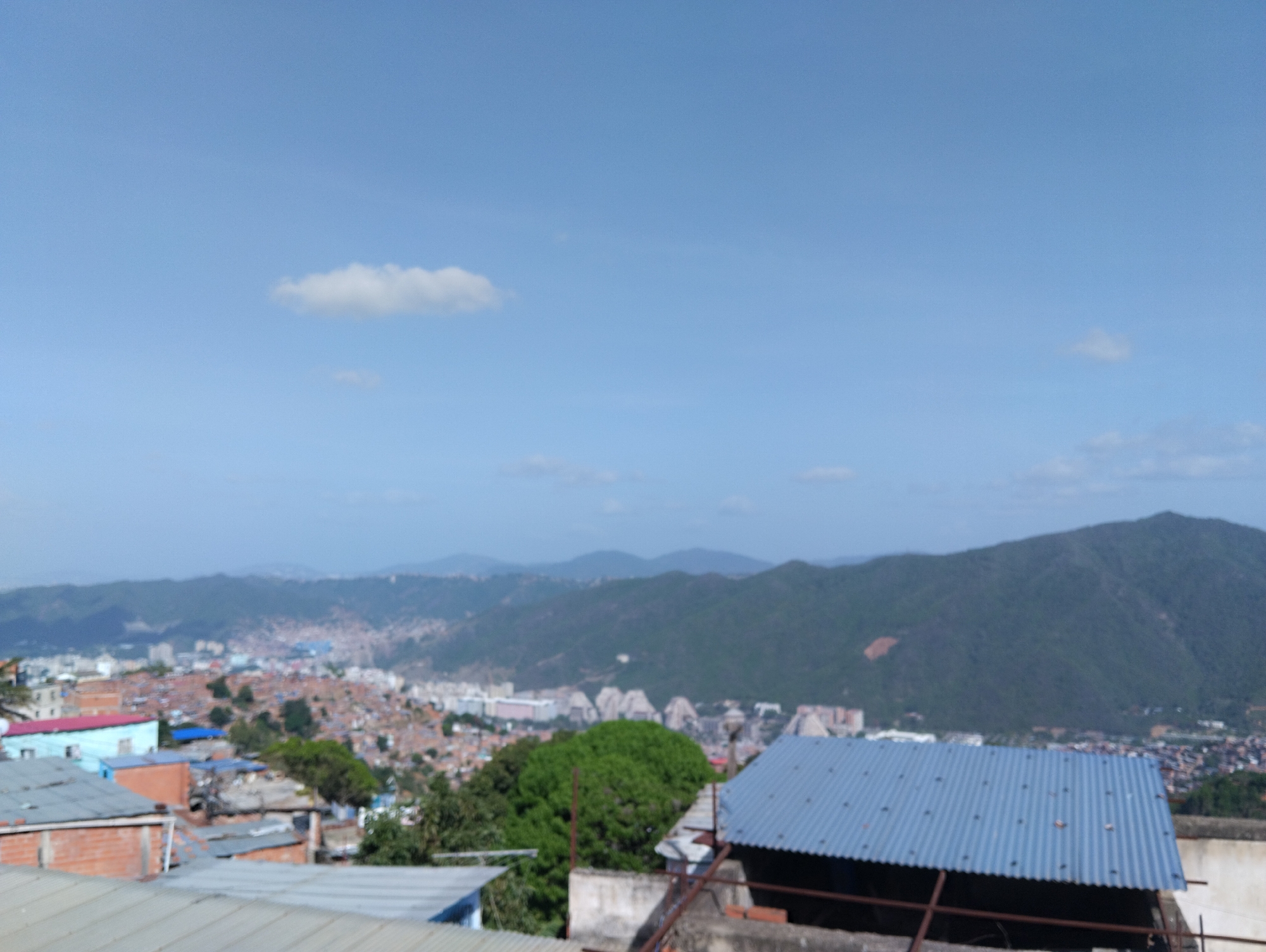 The image shows a wide-angle view of a cityscape in Caracas, Venezuela, with a mountainous backdrop under a partly cloudy blue sky. In the foreground, rooftops of buildings constructed from brick, concrete, and corrugated metal are visible. The midground reveals a densely packed urban area with a mix of informal housing and more structured buildings. The background features verdant mountains.