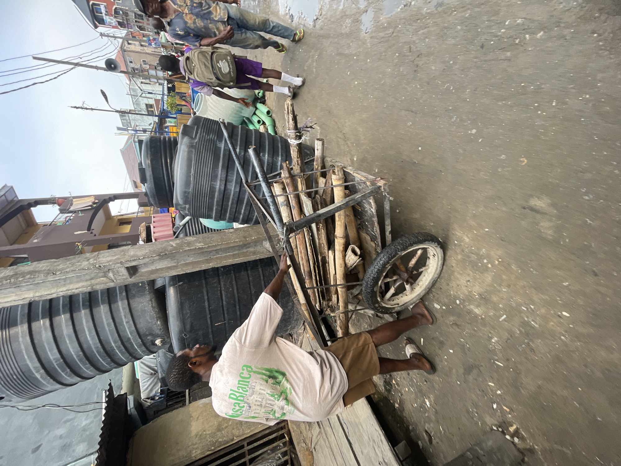 In a street scene in Lagos, Nigeria, a man is pulling a wheeled cart loaded with black plastic water storage tanks. The cart also contains wooden poles. The man is wearing a white t-shirt with "Casa Blanca" printed on the back and brown shorts, with sandals. Other people are seen walking on the street, and buildings are visible in the background. The road surface appears wet and uneven.