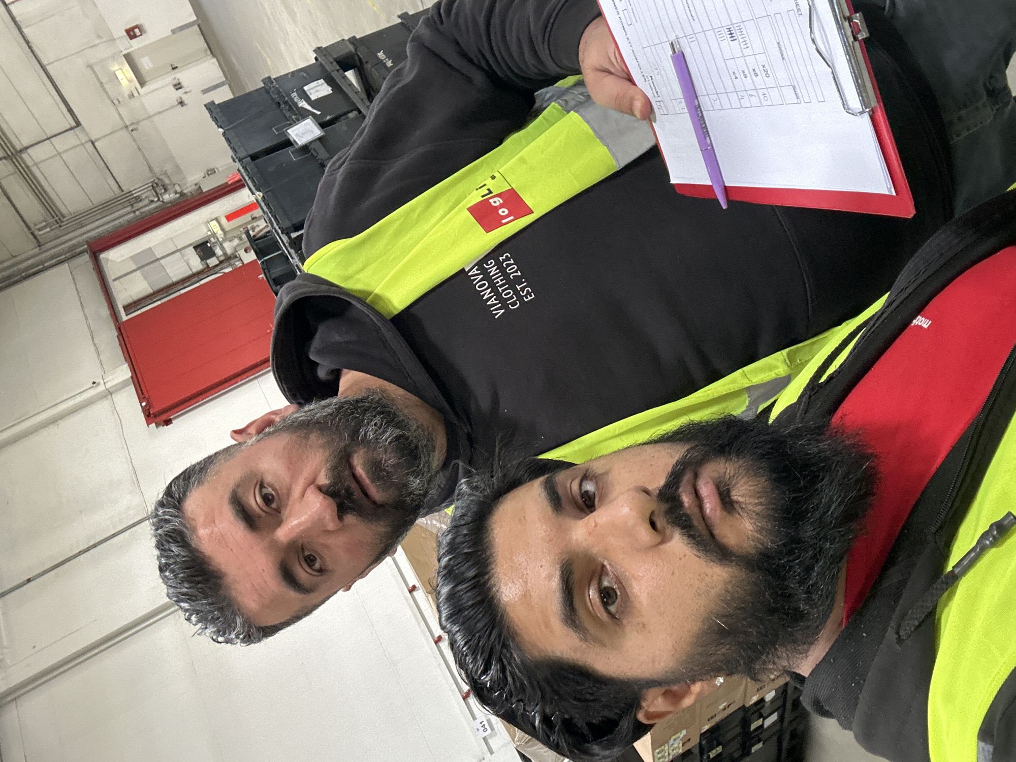 Two men are posing for a selfie in what appears to be a warehouse in Schweitenkirchen, Germany. The man in the foreground, on the left, has dark hair, a beard, and is wearing a red shirt and a high-visibility vest. The man behind him, on the right, has graying dark hair, a beard, and is wearing a black hoodie with a high-visibility vest over it. The hoodie has "VIANOVA CLOTHING EST. 2023" printed on it, along with a red square logo that says "logl i". The man on the right is holding a clipboard with a document and a purple pen. In the background, there are stacks of black plastic crates and a large red door, suggestive of a logistics or distribution center. The overall lighting is bright, indicating an indoor industrial environment.