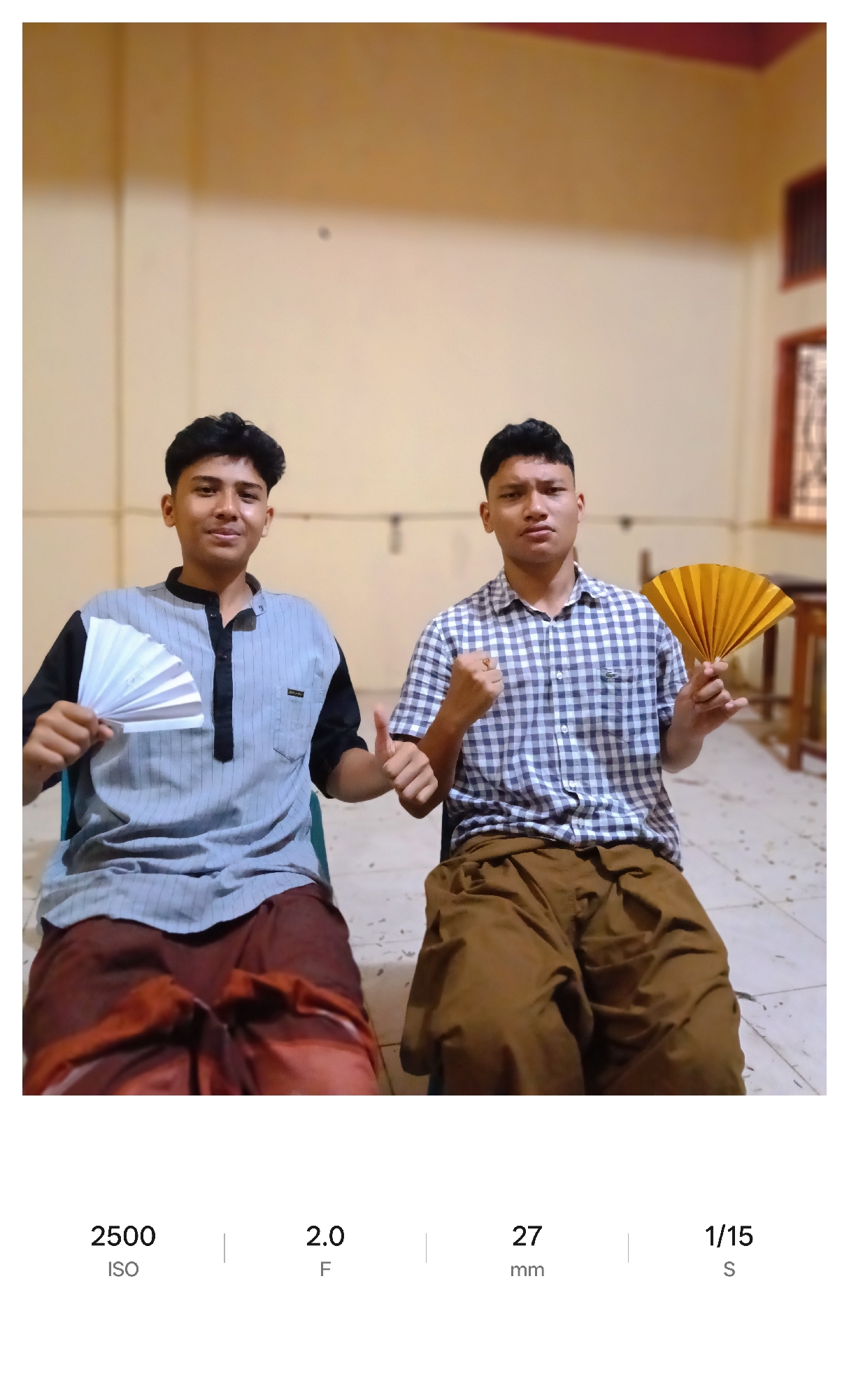 Two young men sit next to each other in an indoor setting, holding small hand fans. The man on the left has dark hair, a gray shirt with black accents, and is holding a white fan. The man on the right has short dark hair, a blue and white checkered shirt, and is holding a yellow fan. Both men are wearing sarongs, brown and maroon. The background shows beige walls and a window with bars. The location is Lhokseumawe, Indonesia.