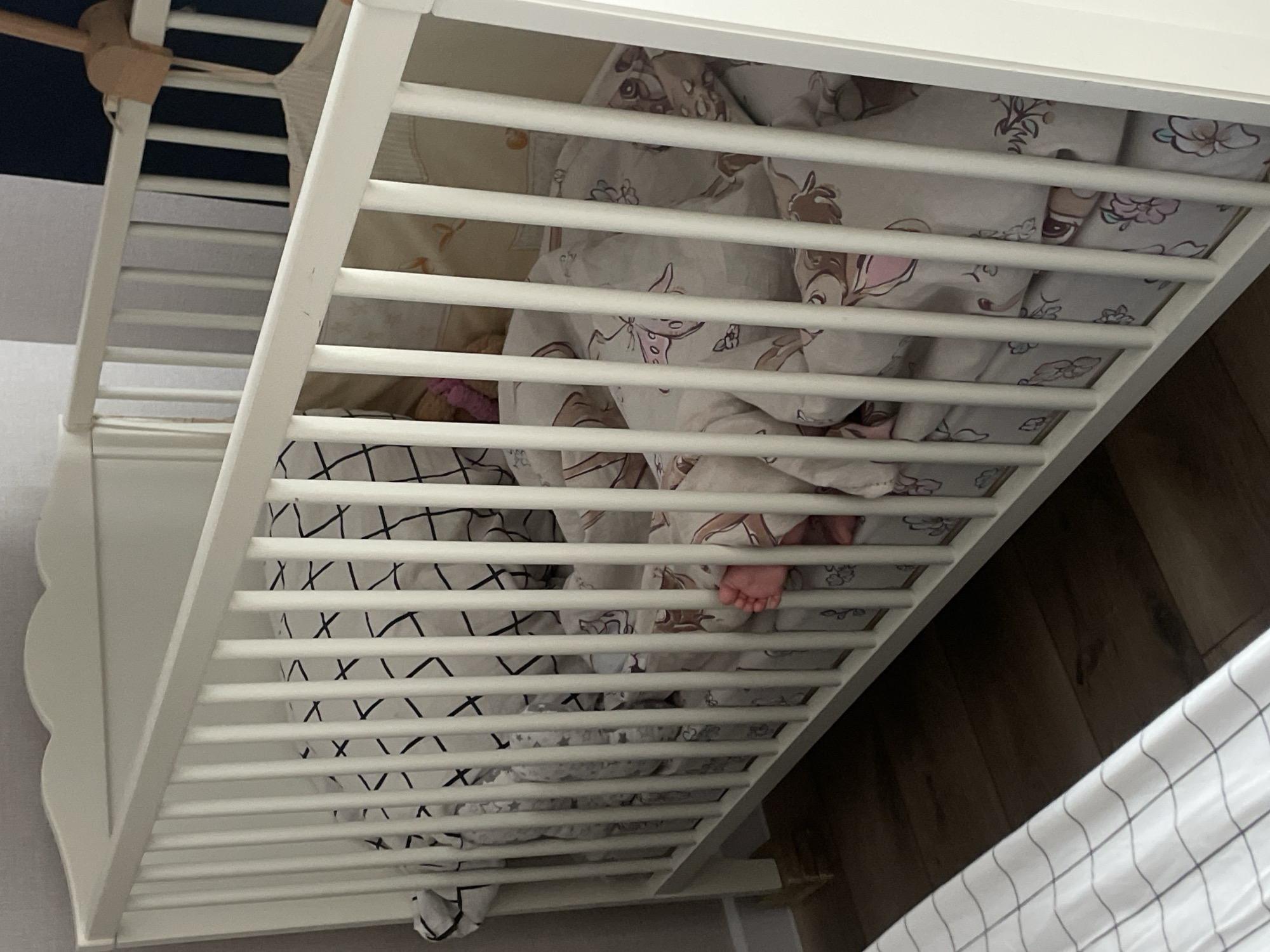 The image captures an overhead, angled view into a white wooden crib, revealing a child sleeping or resting inside. The scene is set indoors, likely in a nursery or bedroom in Podshivalovo, Russia.
The main subjects are the crib and the bedding, with a small baby or child partially visible. The crib features white vertical slats along its sides, and one end panel has a decorative scalloped or wavy edge. A wooden arm, possibly for a baby mobile, extends from the top left corner of the crib.
Inside the crib, multiple layers of bedding are present. A sheet or blanket with a black irregular grid pattern is visible on the left side and beneath other covers. Over this, a light-colored duvet or blanket adorned with a pattern of cartoon deer (reminiscent of Bambi) and floral motifs in pink and brown tones is draped. A tiny, bare baby foot is seen peeking out from under these blankets through the crib slats on the right side, indicating the child's presence.
The surrounding environment includes a dark blue or navy wall visible above the crib on the left, and a wooden floor visible on the right side beneath the crib. The lighting appears to be ambient indoor light. No text is visible in the image.