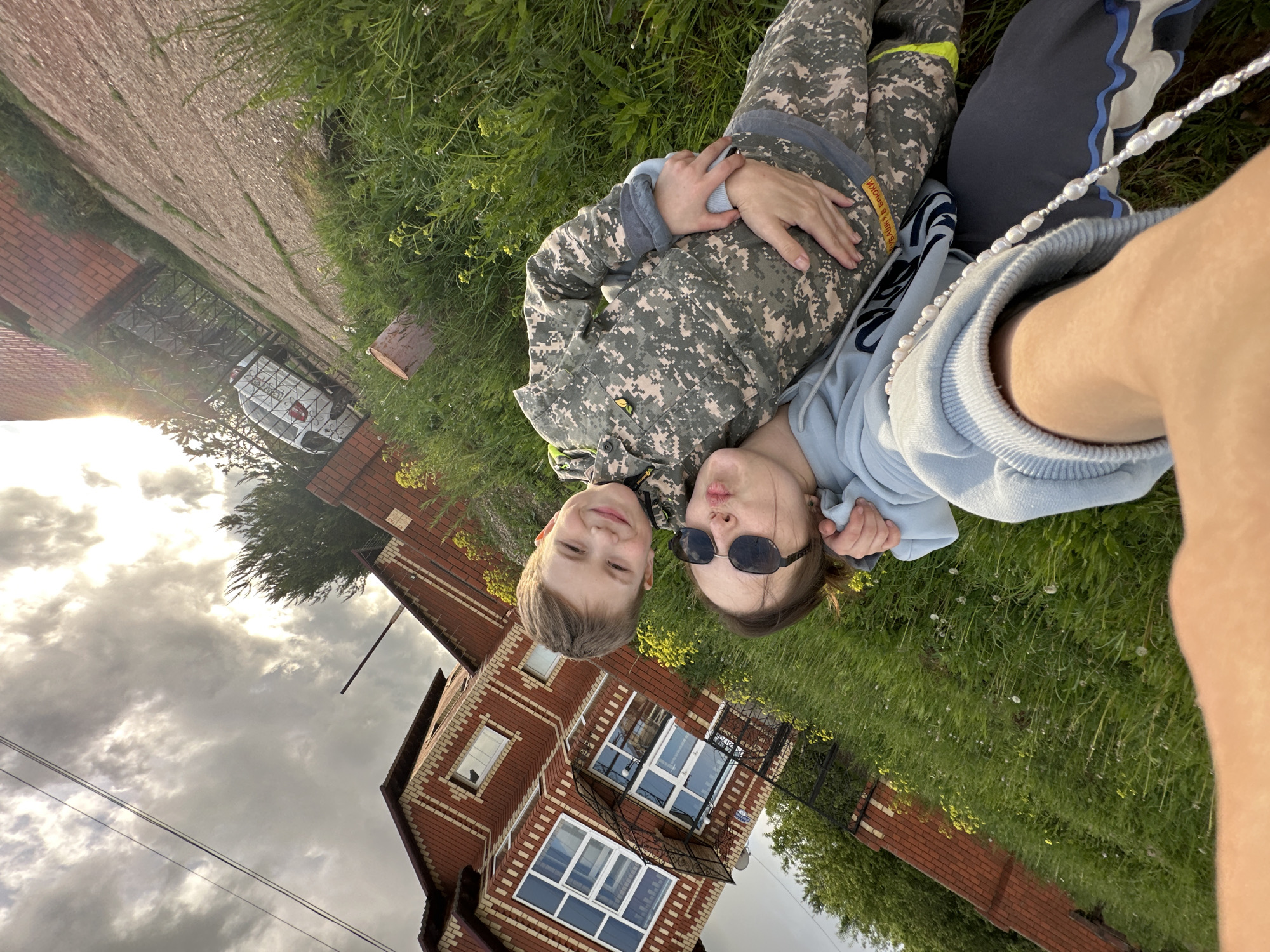 Two children, a boy and a girl, are lying on their backs in a grassy area. The boy is wearing camouflage clothing. The girl is wearing a light blue sweatshirt and sunglasses. They are positioned close together, appearing to interact for a selfie.
Behind them is a two-story brick house with white-framed windows and a wrought-iron balcony. The house has a brick exterior. Next to the house is a paved path. A vehicle is behind a fence.
The location is Ilyinsky, Russia. The sky is overcast.