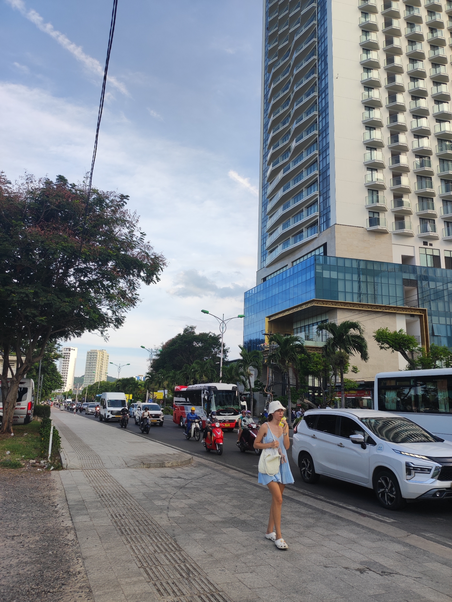 In Nha Trang Ward, Vietnam, a young woman walks along a busy sidewalk next to a street congested with traffic. She is wearing a light blue sundress, a white bucket hat, and white slip-on shoes, and carries a woven white bag. She is holding a yellow object to her mouth, possibly a drink or snack. The street is filled with cars, buses, and numerous motorbikes. A white minivan is parked to her left, and further down the street, a large white bus is visible. A tall, modern building with a white facade and balconies dominates the background, its reflective windows showing a distorted image of the sky and clouds. Other buildings and trees, including palm trees, are also visible. The sky is partly cloudy, suggesting it is daytime, likely late afternoon or early evening due to the soft light. The overall scene depicts a typical urban environment with moderate traffic and pedestrian activity.