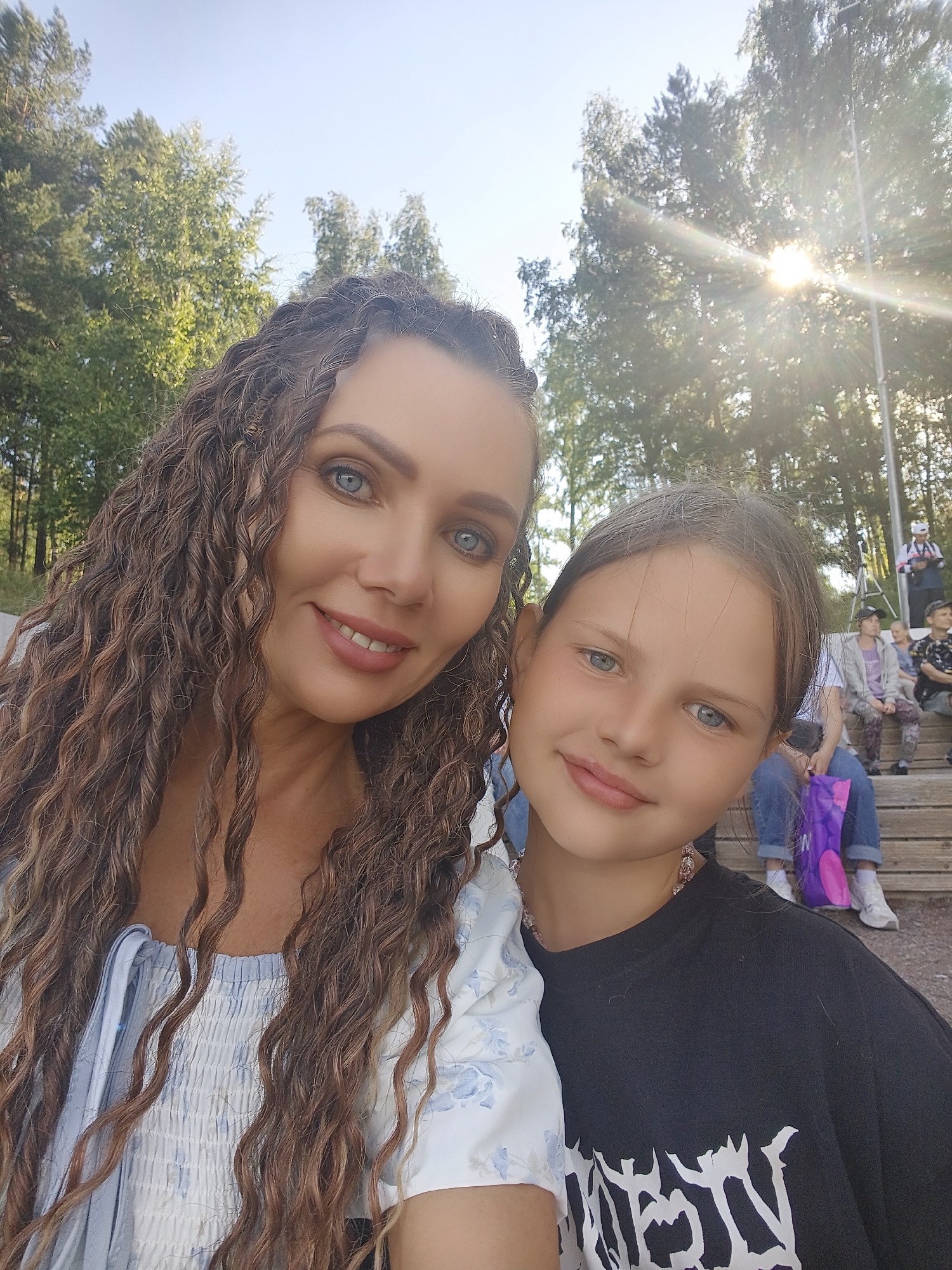 A selfie taken outdoors shows two females. The female on the left has long, curly brown hair and is smiling. She is wearing a white and blue top. The female on the right has long brown hair and is also smiling. She is wearing a black t-shirt. Both are positioned close to the camera. In the background, there are trees, a clear blue sky, and a bright sun with visible rays. Wooden bleachers are visible behind the females, with several other people seated on them. The overall impression is a sunny day, possibly at an outdoor event or gathering. The location is not specified in the prompt.