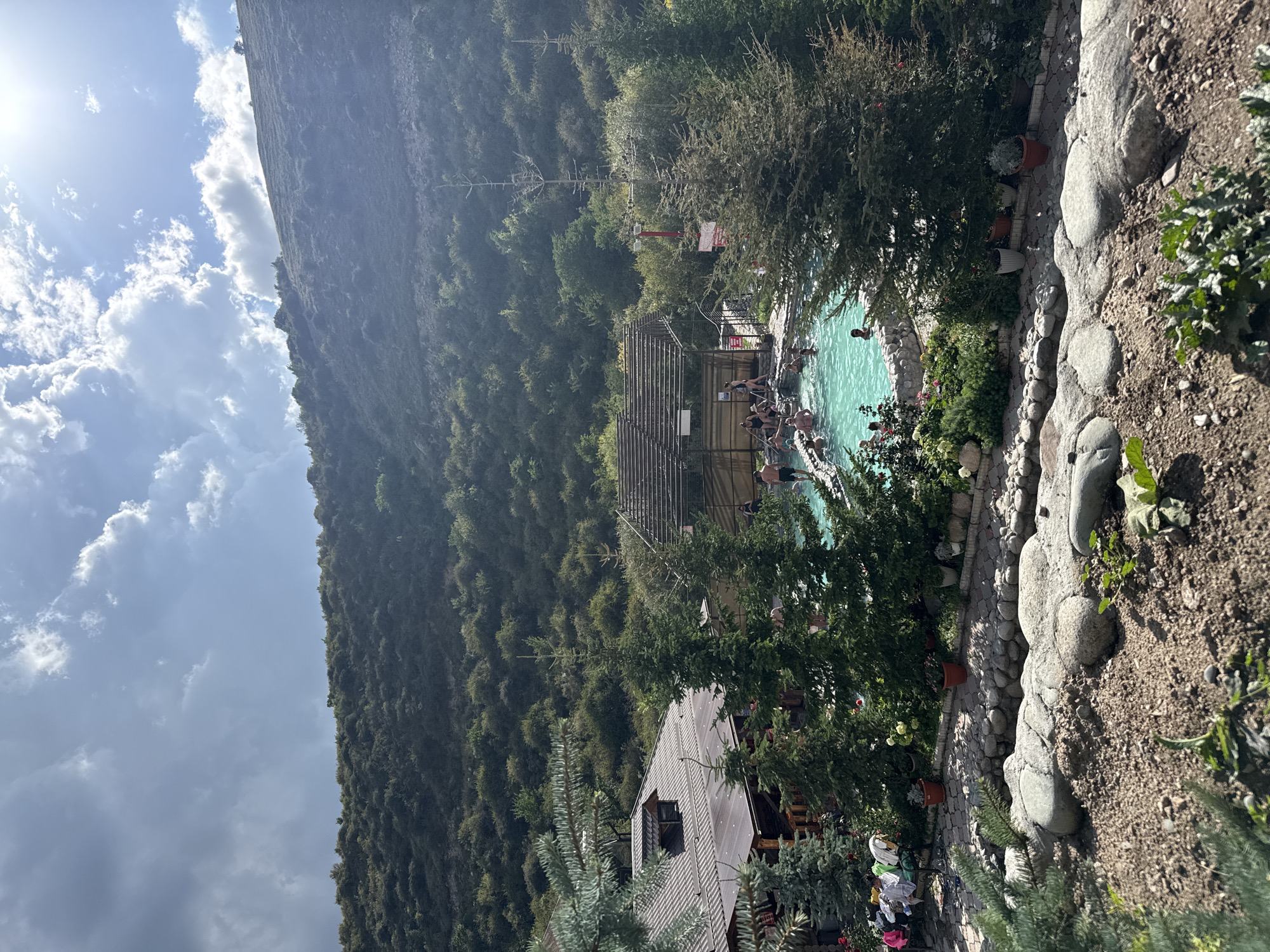 In Ak-Suu, Kyrgyzstan, a natural hot spring resort is depicted. The image shows a vibrant turquoise pool filled with people enjoying the water. The resort features rustic wooden buildings with sloped roofs, surrounded by lush green trees and a hillside covered in dense vegetation. In the foreground, a landscaped area with large rocks and gravel adds to the natural ambiance. The sky is partly cloudy, with the sun casting a bright light.