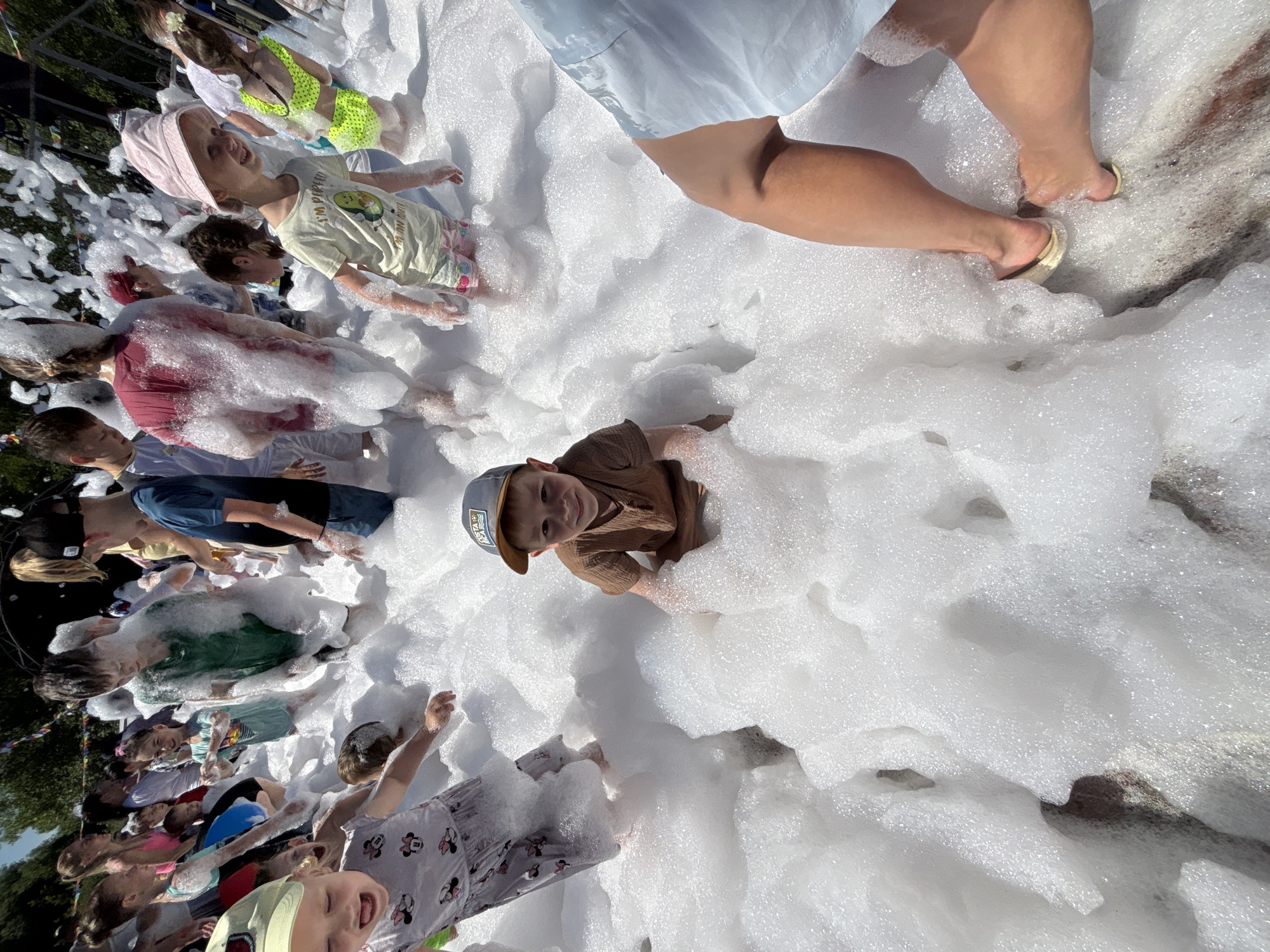 This is a high-angle, full shot of children playing in a foam party. The location is a public outdoor area, likely a park or festival ground, in Zavolzhye, Russia, judging by the presence of a Cyrillic text on a child's hat. The ground is covered in a thick layer of white foam, obscuring most of the lower bodies of the children.
In the foreground, a young boy wearing a brown t-shirt and a grey baseball cap with "COSTA RICA" visible on it, is kneeling in the foam and looking directly at the camera. To his left, a young girl in a grey dress with a Mickey Mouse pattern is partially visible, also playing in the foam. In the mid-ground and background, a crowd of children of various ages are immersed in the foam, some actively playing and others standing. Their clothing is mostly casual summer attire. A few adults are present at the edges of the frame, with their legs visible. The environment appears to be sunny with some trees visible in the background, suggesting an outdoor event during daytime. The overall atmosphere is one of playful enjoyment and summer fun.