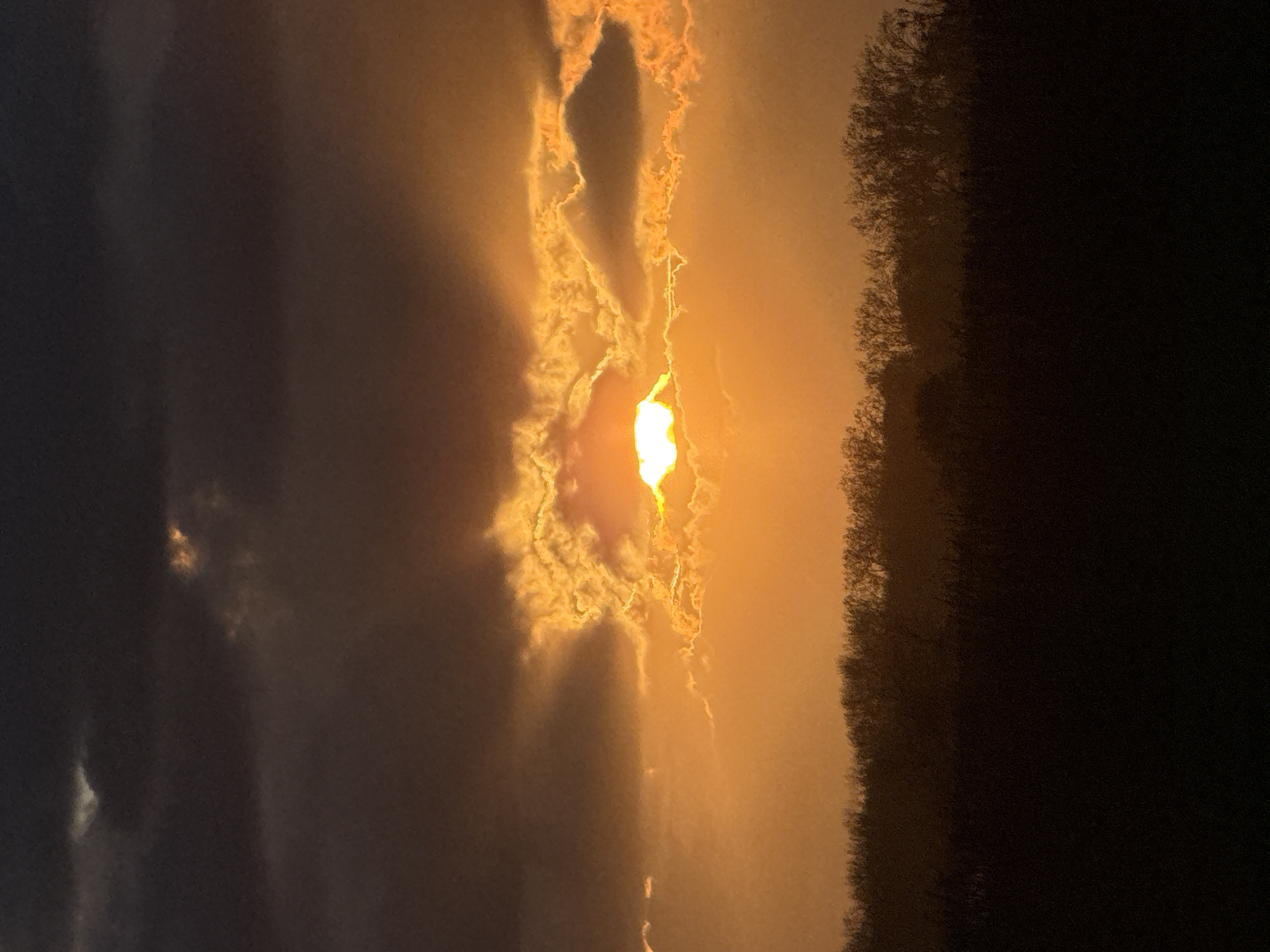 The image depicts a sunset or sunrise scene, likely taken in Gorleston-on-Sea, United Kingdom. A bright, intense light source, presumably the sun, shines through a gap in the clouds. The clouds surrounding the light source are illuminated with a golden hue. To the right, a dark mass suggests a treeline or dense vegetation. The sky darkens towards the left of the frame. The image is vertically oriented.