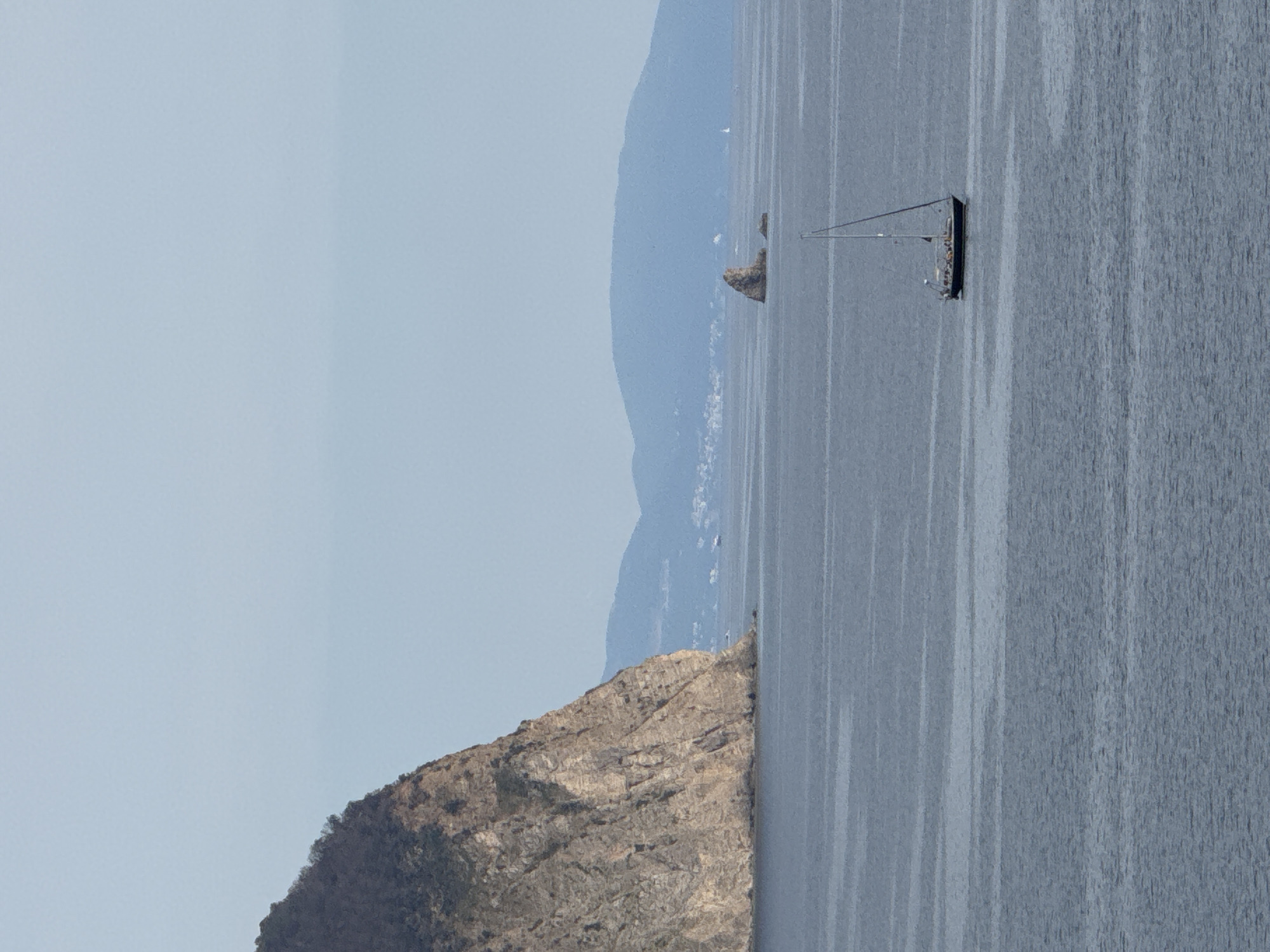 The image depicts a serene seascape with a large, rocky cliff face on the left, sparsely covered with vegetation. The cliff dominates the left side of the frame, its textured surface showing layers and crevices. In the foreground, the calm, deep blue water of the sea stretches across the image. A solitary sailboat with a single mast is visible in the middle right of the frame, its dark hull contrasting with the lighter sea. In the distance, a series of hazy blue mountains form the horizon, with a small, rugged rock formation rising from the sea between the mountains and the sailboat. The sky above is a pale, overcast blue, with no visible sun. The overall atmosphere is tranquil and natural, suggesting a coastal landscape, potentially in Mirište, Montenegro, given the geological features and marine environment.