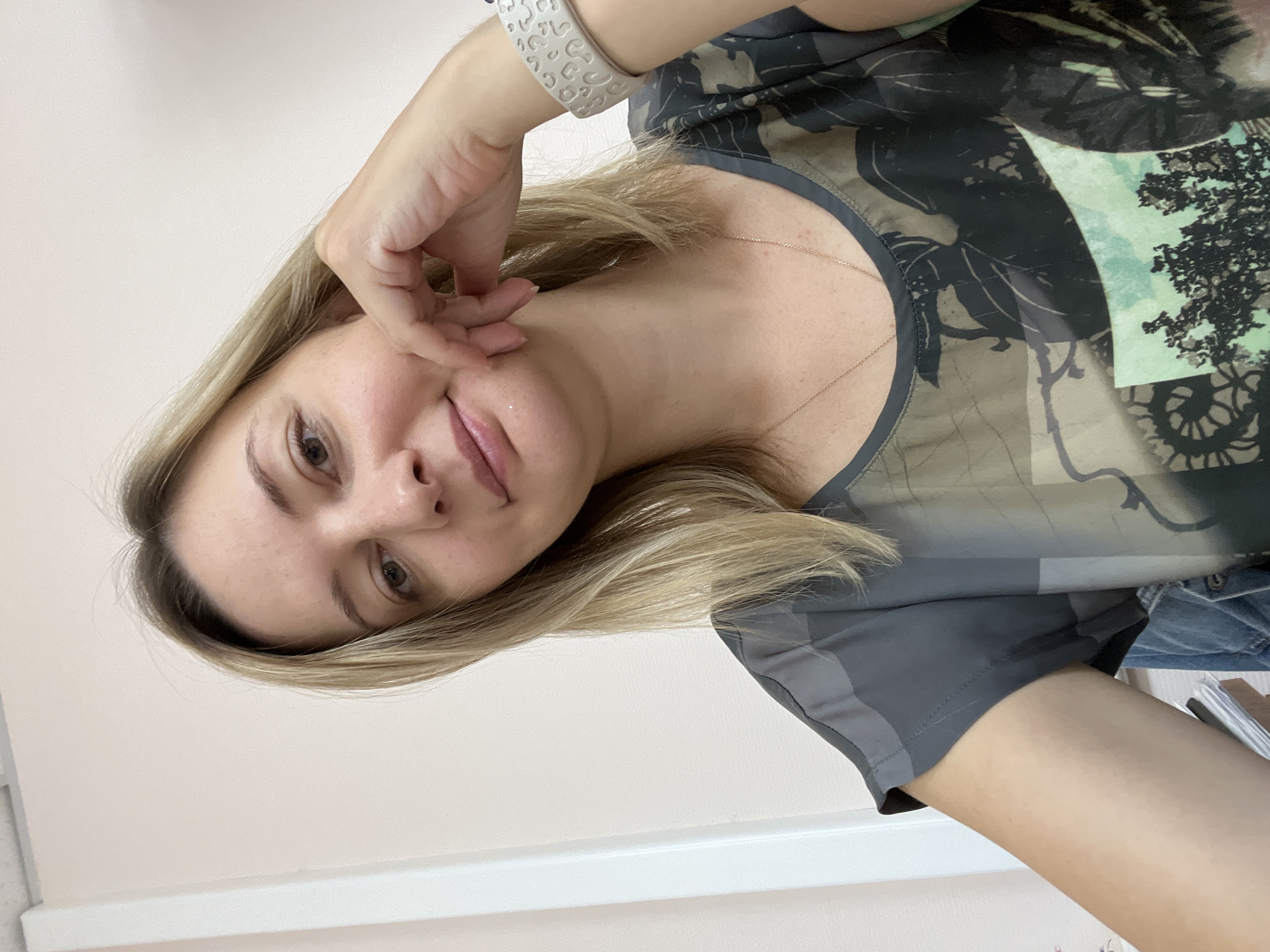 A medium shot image shows a woman with blonde hair and fair skin, facing the camera and resting her right hand on her cheek. She is wearing a dark-colored shirt with a graphic print featuring leaves and trees. A silver-colored bracelet with a patterned design is visible on her right wrist. The background is a plain, light-colored wall with a white vertical structure on the left. The lighting appears to be indoor, with no indication of an outdoor environment or a specific city. The image does not provide any information to determine the location in Volgograd, Russia.