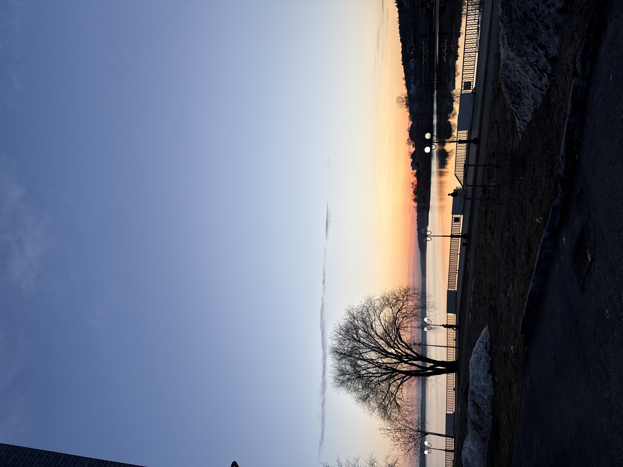 This image captures a serene waterfront scene in Votkinsk, Russia, likely during sunrise or sunset. The sky displays a beautiful gradient, fading from a clear, deep blue overhead to soft orange, pink, and yellow tones at the horizon, indicating the low angle of the sun. These warm colors are mirrored in the calm waters of what appears to be a large pond or reservoir, creating a luminous reflection.
In the foreground, a paved promenade stretches along the water's edge, bordered by a decorative metal railing. Several classic-style streetlights with two globes each are illuminated, casting warm glows on the path and water, suggesting the light is fading or just beginning to appear. Bare deciduous trees, including a large, prominent one in the mid-ground with intricate branches, stand sentinel along the embankment. Patches of white snow or ice are visible on the grassy areas beside the path, suggesting a cold season like late winter or early spring. The distant bank across the water is dark and appears to be covered with forested hills or sparsely populated land with some scattered buildings.
The overall scene is devoid of people or visible activity, conveying a sense of peace and quietude. There is no discernible text visible in the image.