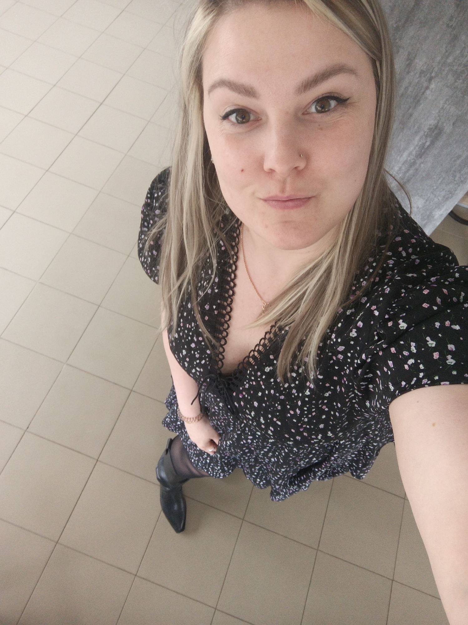 A woman is taking a selfie from a high-angle perspective. She has blonde hair, fair skin, and is wearing a black dress with a floral pattern and lace trim. She is also wearing dark tights and black shoes. She has a nose piercing and a gold necklace. The background consists of tiled flooring and a grey surface. The image appears to have been taken indoors. The location details are not visible in the image.