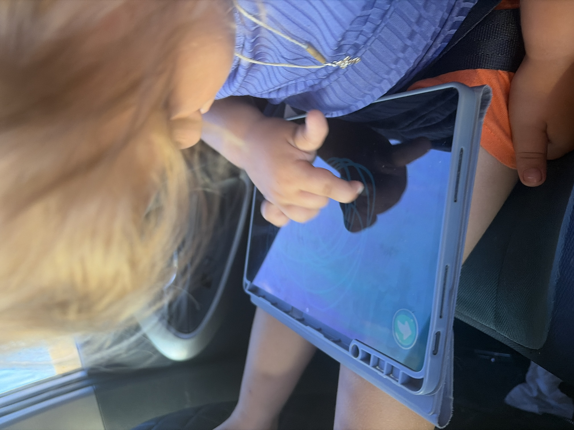 A young child, with blonde hair, is sitting in a car and using a tablet. The child is wearing a purple shirt and orange shorts. The tablet is in a blue case and the screen is displaying a colorful app. The child's left hand is resting on the tablet, and their right index finger is touching the screen. The background is blurred, but it appears to be the interior of a car. The scene is brightly lit, likely by sunlight coming through the car windows. The image quality is slightly blurry, but the details are still discernible.