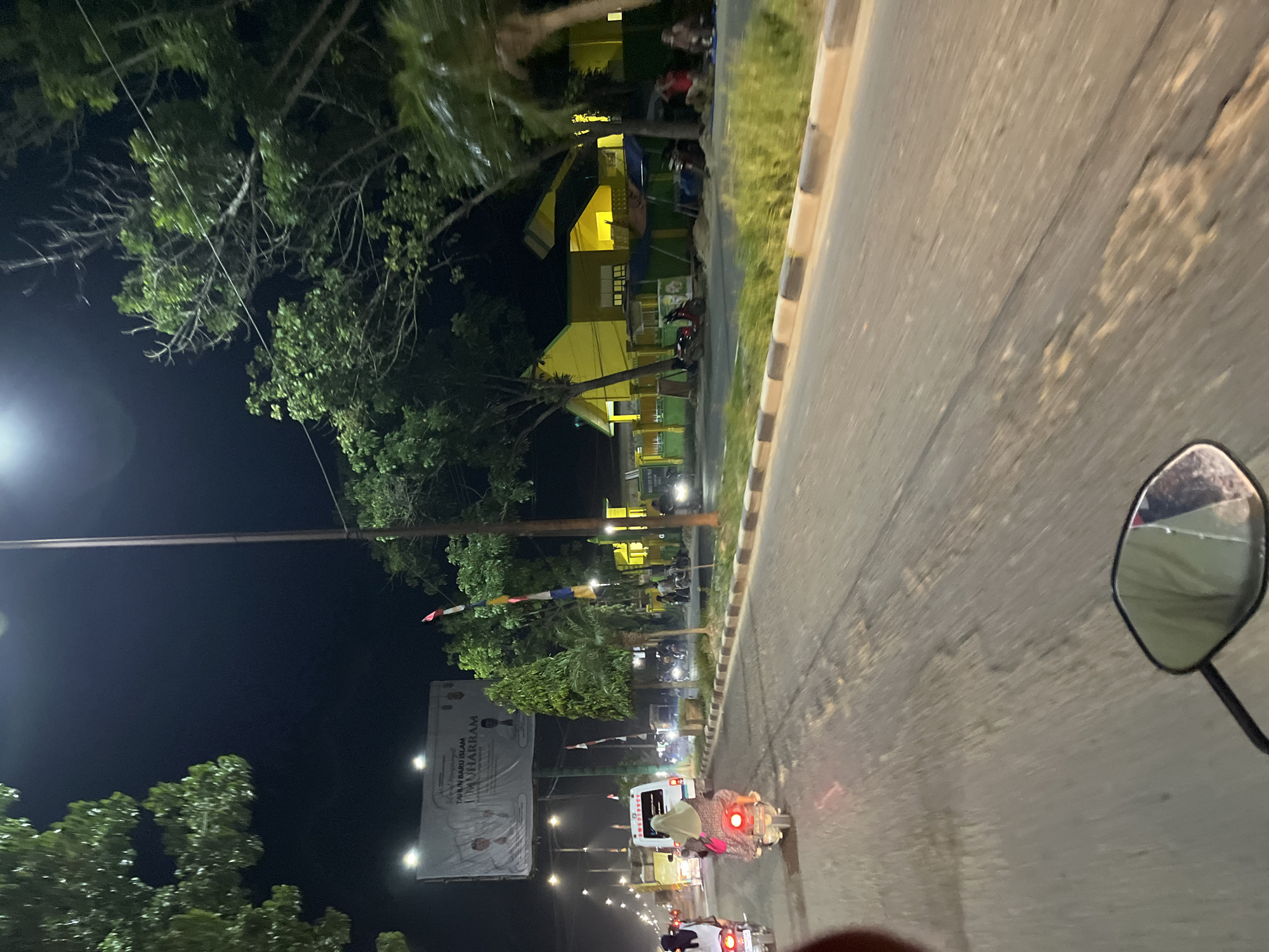 The image captures a nighttime scene on a street in Bundar, Indonesia, as seen from the perspective of a motorcycle rider. The road is paved and shows signs of wear, with some patches and cracks. To the right of the road is a grassy area bordered by a concrete curb.
On the left side of the road, a brightly lit yellow building with green accents is visible, possibly a school or a public facility. Trees line the street, their leaves illuminated by streetlights and the building's lights, creating shadows on the road. Small flags, possibly red and white, are strung between poles, adding a touch of color to the scene.
Several vehicles are present on the road. In the foreground, the rear of a motorcycle is visible, its taillight glowing red. Further down the road, another motorcycle with a rider wearing a headscarf is visible, also with its taillight illuminated. Other vehicles and motorcycles are scattered along the street, some parked and others in motion. A large banner or billboard is also visible on the left side of the road, displaying text that includes "TAHUN BARU ISLAM MUHARRAM".
The time of day is clearly night, indicated by the dark sky and artificial lighting. The weather appears calm, with no signs of rain or strong winds. The overall atmosphere suggests a typical evening in a town or city in Indonesia, with people out and about.