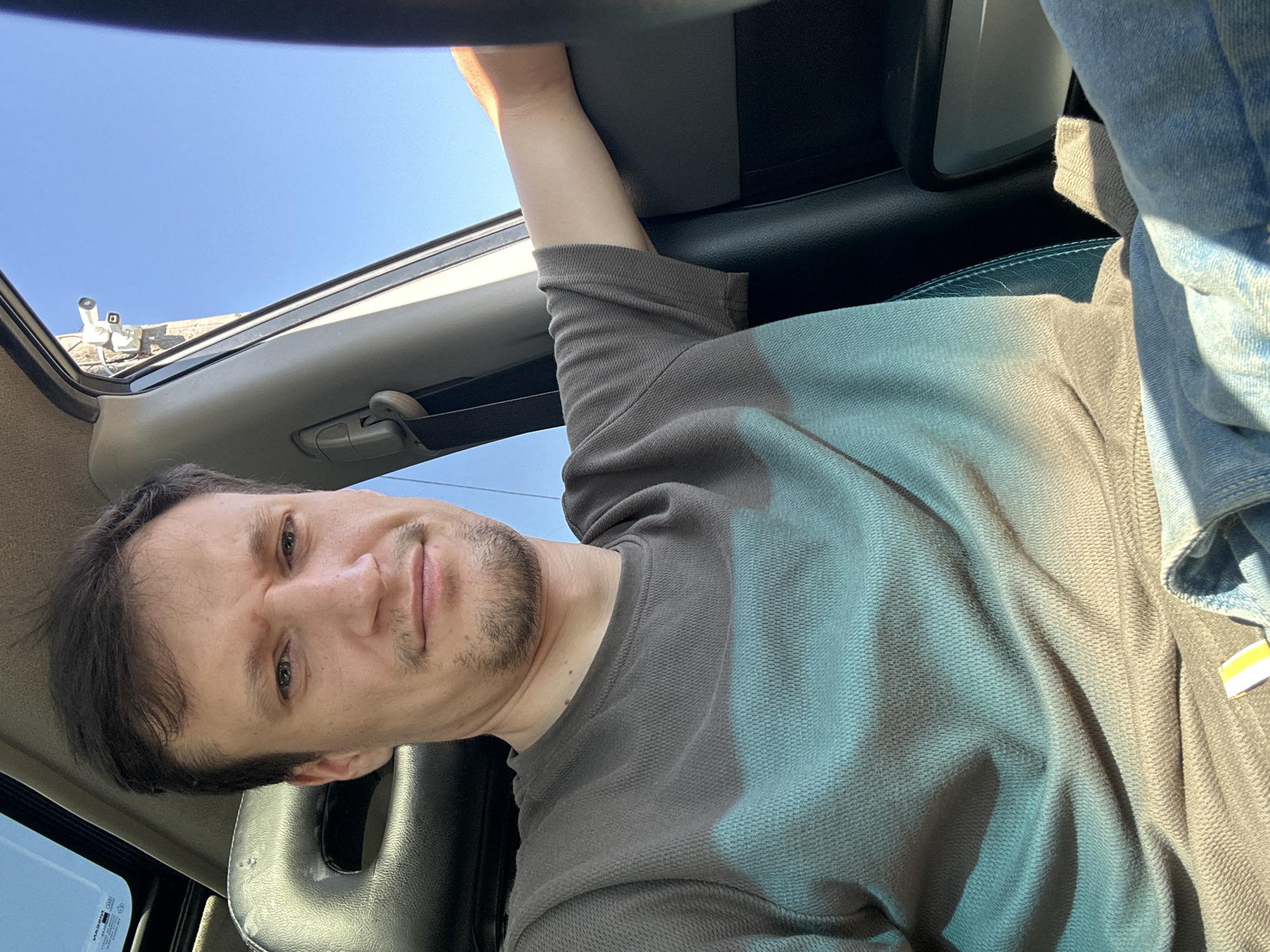 A man is seated in the driver's seat of a car, looking directly at the camera. The car appears to be parked outdoors, with a clear blue sky visible through the window. A security camera is mounted on a wall outside the car, visible through the side window. The man is wearing a gray and teal t-shirt and blue jeans. His right arm is resting on the car door. The image is taken from a slightly elevated angle. The location provided, Shatava, Ukraine, is not depicted in the image.