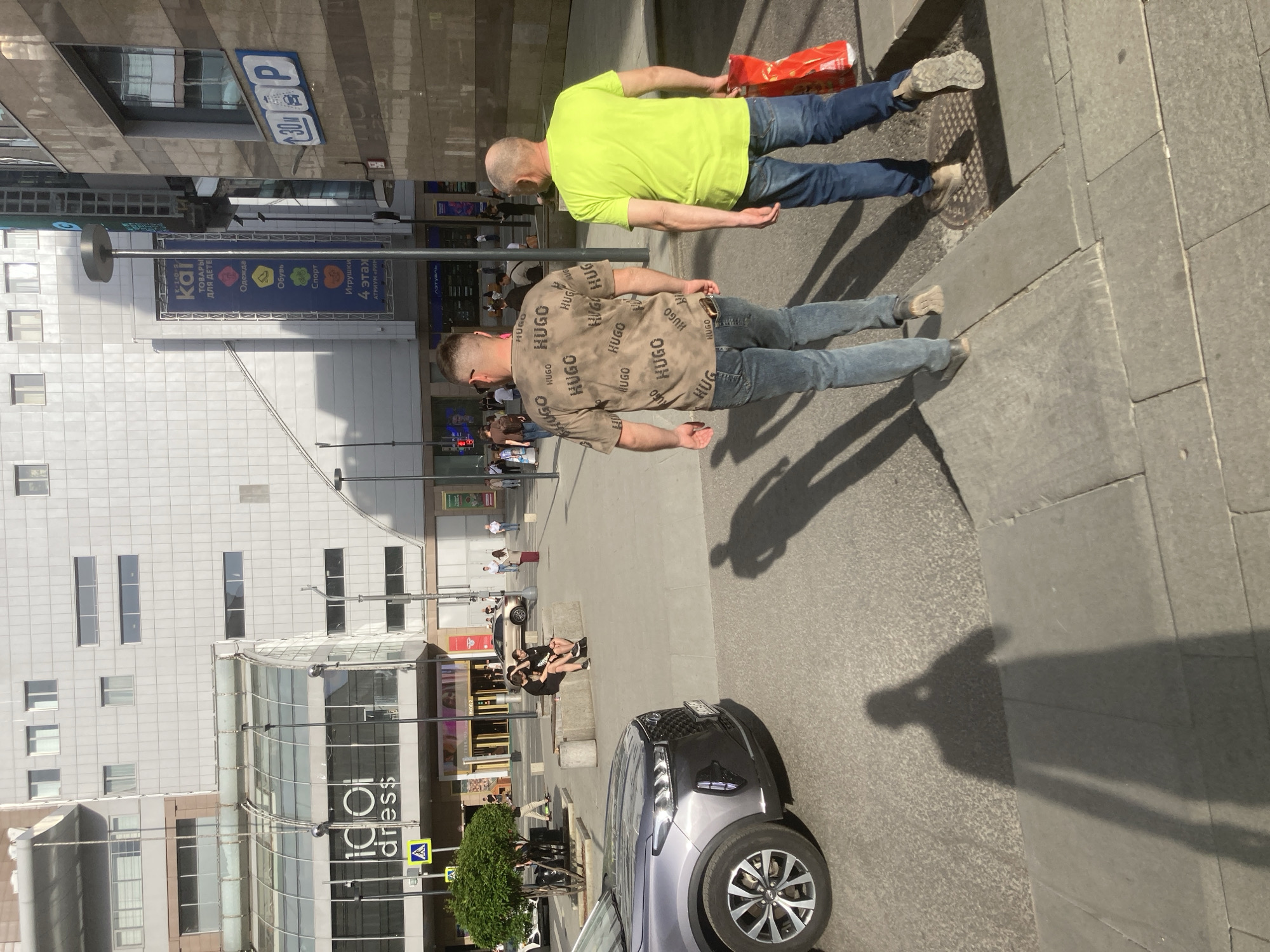 Here's a detailed description of the image:
**Scene:** The image is taken outdoors, likely in a public area within Moscow, Russia. The architecture suggests a modern urban environment.
**People:**
* Two men are walking side-by-side. One is wearing a lime green t-shirt and jeans, carrying a red package. The second is wearing a beige t-shirt with the word "HUGO" printed all over it and jeans.
**Objects:**
* A silver SUV is parked on the street.
* Street signs are visible, with Cyrillic text, which indicates that this is Russia.
* Manhole Cover
* A building with a storefront named "1001 dress" is visible.
* Several commercial signs are placed above. The one with "ТОВАРЫ ДЛЯ ДЕТЕ" indicates this is a shop with goods for kids.
* A light post is centrally positioned.
**Actions/Interactions:**
* The two men appear to be walking together down the street.
* Other people are seen sitting on concrete blocks and walking along the sidewalks in the background.
**Environment:**
* The lighting suggests it is daytime.
* Shadows indicate a sunny day.
* The presence of cars and pedestrians indicates an urban street.
* The buildings are modern and made from glass and concrete, which are common elements in city architecture.