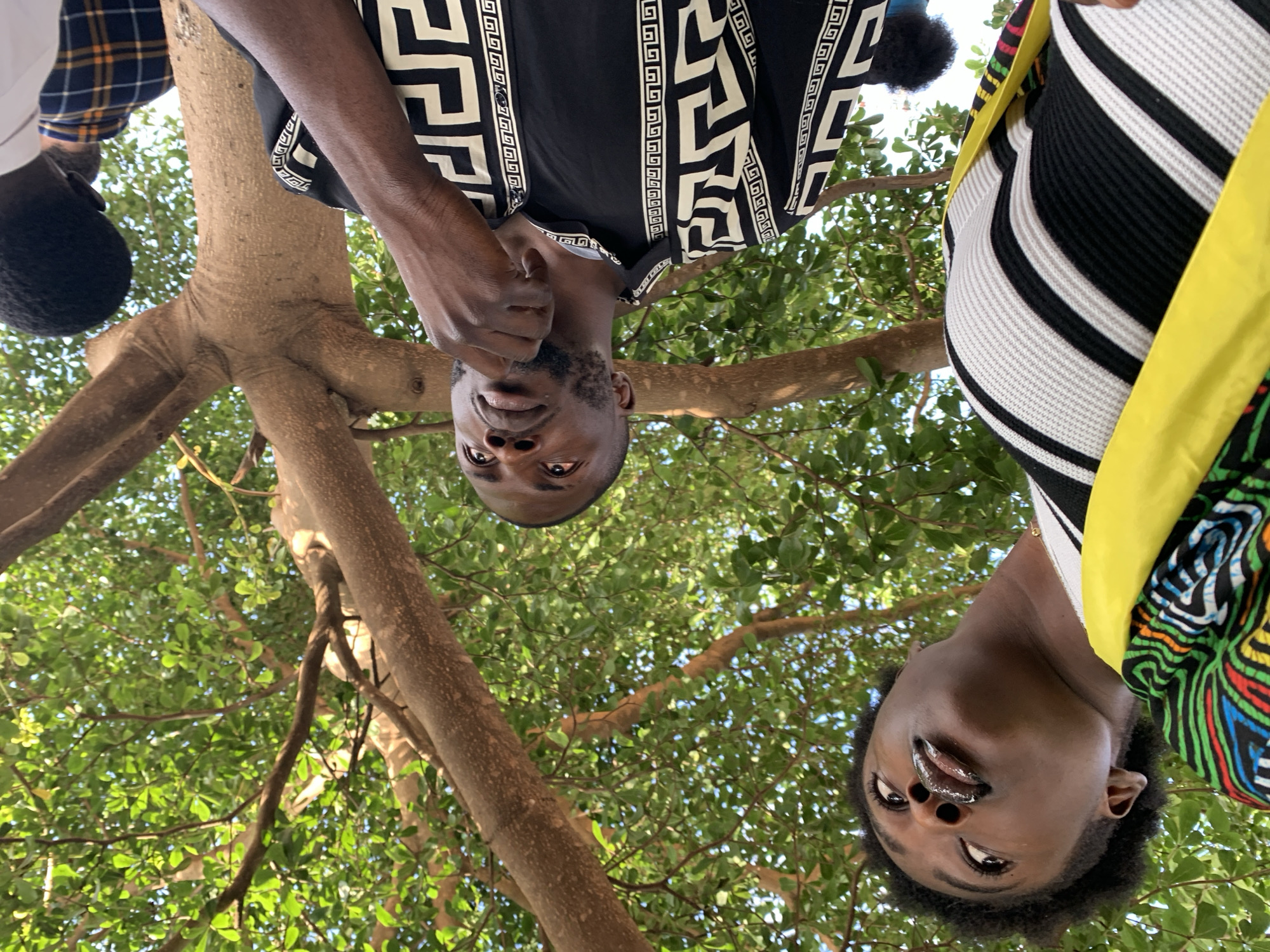 This is a low-angle, waist-up shot of two individuals, a man and a woman, standing outdoors under a tree. The man is on the right, wearing a black shirt with a white geometric pattern, and his hand is near his chin. The woman is on the left, looking upwards, wearing a striped shirt and a yellow accessory. Both individuals have dark skin tones. The background consists of the branches and green leaves of a tree, with hints of a blue sky visible. The image appears to be taken in Rafin Par, Nigeria.