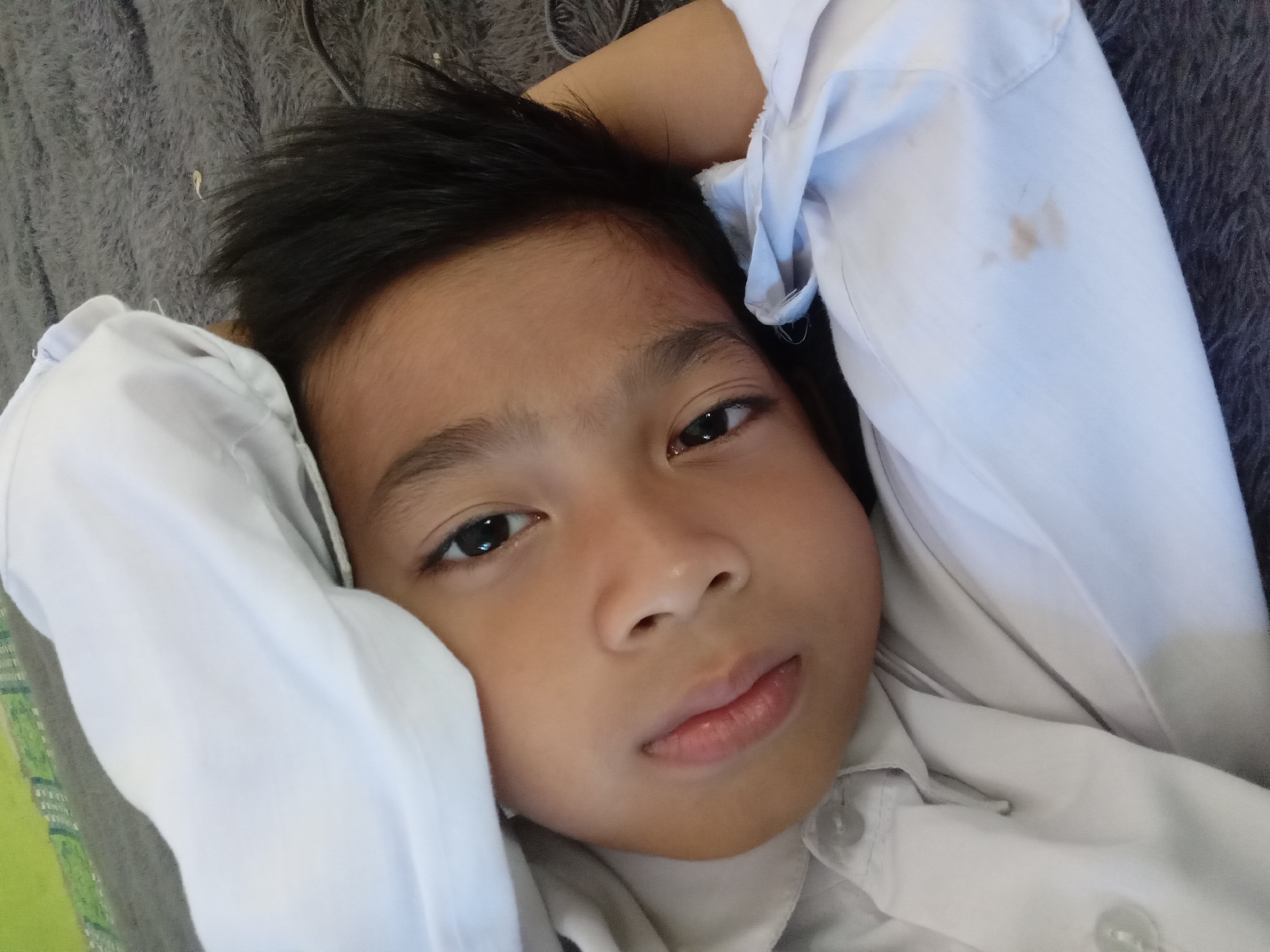 The image shows a close-up, low-angle shot of a young child lying on a gray, furry surface, possibly a carpet or blanket. The child is wearing a white collared shirt with visible buttons. Part of the child's white shirt is pulled over their head, obscuring the top of their head. The child's face is turned towards the camera, with their right hand resting behind their head. Their left arm is bent and partially visible. The child's eyes are open and looking directly at the camera. The overall lighting suggests an indoor setting, likely daytime. The presence of a patterned green object in the upper left corner of the image might indicate a mat or part of the background. There are a few small, light brown stains on the child's white shirt. The image composition is tight, focusing primarily on the child's face and upper body.