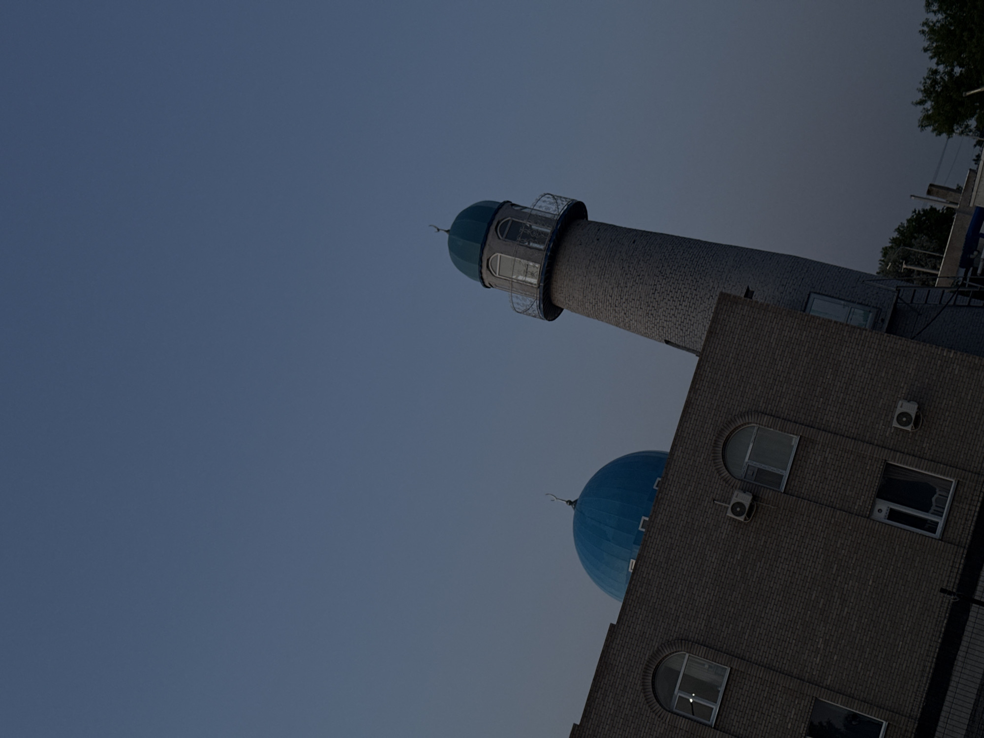 A low-angle shot presents the minaret and dome of a brick building, possibly a mosque, against a dusk sky. The minaret, cylindrical in shape and constructed of light-colored bricks, tapers towards the top, culminating in a blue dome and a viewing platform with a railing. The building's facade, also brick, includes arched windows, standard rectangular windows, and air conditioning units. The angle and the dim lighting obscure specific details, but the architectural elements are clearly visible. The absence of people and context leaves the building's function and exact location unspecified.