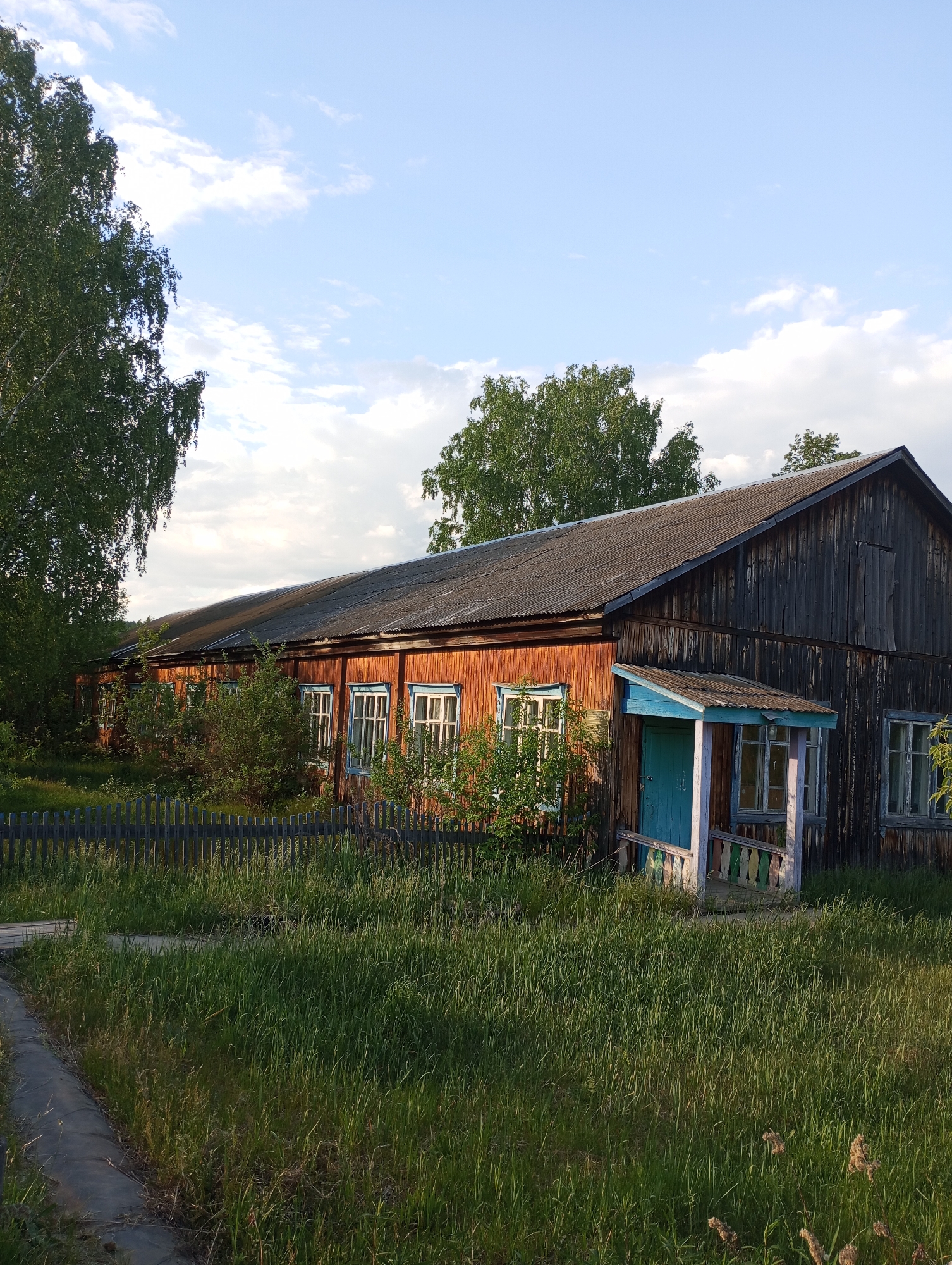 The image shows a long, one-story building in Буренка, Russia. The building's structure appears to be mainly wooden, with the right side noticeably darker and weathered. The middle section has reddish-brown siding. Windows with light blue frames are distributed along the middle section. A small, turquoise-colored porch with a roof is attached to the right side of the building. Tall grass dominates the foreground. A dark gray wooden fence runs along the front of the building, and there is a concrete path on the left side of the frame. Several trees surround the building. The sky is blue with scattered clouds.