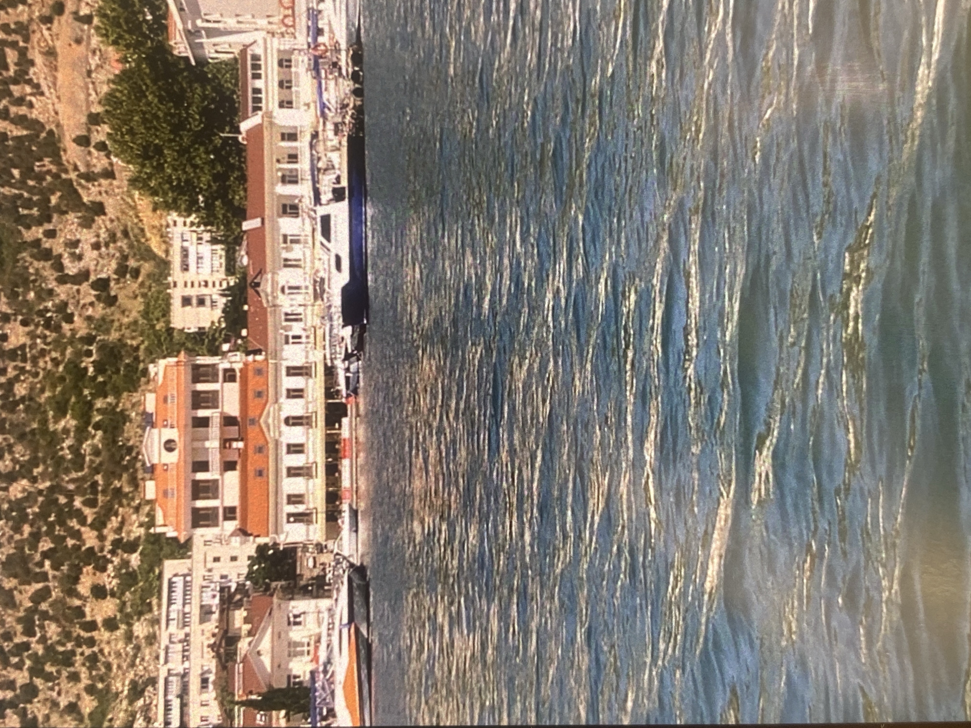 A wide-angle, eye-level medium shot captures a sunlit harbor with a backdrop of coastal buildings and a rocky, tree-covered hillside under a clear sky. The foreground is dominated by the choppy, sun-dappled surface of the sea, with waves creating a shimmering texture.
In the middle ground, several boats are docked or moored along a waterfront. The most prominent is a sleek, dark-hulled motorboat with a white cabin, positioned towards the right side. To its left, a white vessel with an orange stripe is visible. Beyond these boats, a line of low-rise buildings with red-tiled roofs and white facades runs along the water's edge. These buildings appear to be shops or restaurants, with awnings and open windows visible.
Further inland, a larger, multi-story building with a similar red-tiled roof and white facade stands out. This building has multiple balconies and arched windows, suggesting it might be a hotel or residential complex. Behind this, a cluster of lighter-colored buildings, possibly apartment blocks, are visible against the hillside. The hillside itself is covered in dense green vegetation and appears rocky, typical of a Mediterranean or similar coastal landscape. The overall impression is a vibrant, active harbor scene in a picturesque coastal town.