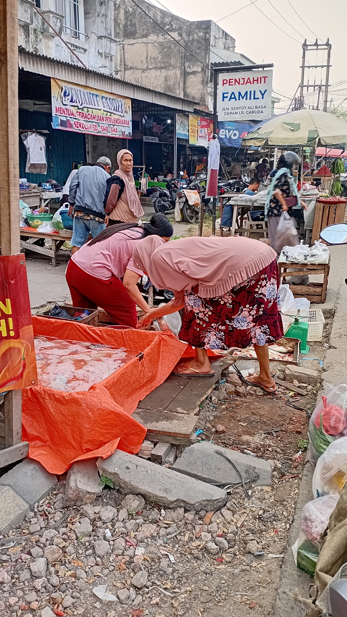 The image depicts an outdoor market scene, likely in the city of Jambi, Indonesia. In the foreground, two individuals are actively engaged with a large orange tarp-lined container filled with what appears to be fish. One person, wearing a pink top and red pants, is bending over the container. The other person, in a pink top and a floral skirt, is also leaning into the container, with their hands submerged. They are both wearing headscarves. A third person, a man in a blue jacket and jeans, stands nearby and looks towards the viewer. The background shows a street lined with various market stalls and shops. A sign above one shop reads "PENJAHIT FAMILY" (Family Tailor), with an address below it indicating "JLN. SENTOT ALI BASA DEPAN LR. GARUDA" in Jambi. Motorcycles are parked in front of some of the stalls, and other market-goers are visible in the distance. The ground is paved with concrete and dirt, with debris scattered about, suggesting a busy and somewhat unkempt environment. The overall atmosphere is that of a typical bustling local market.