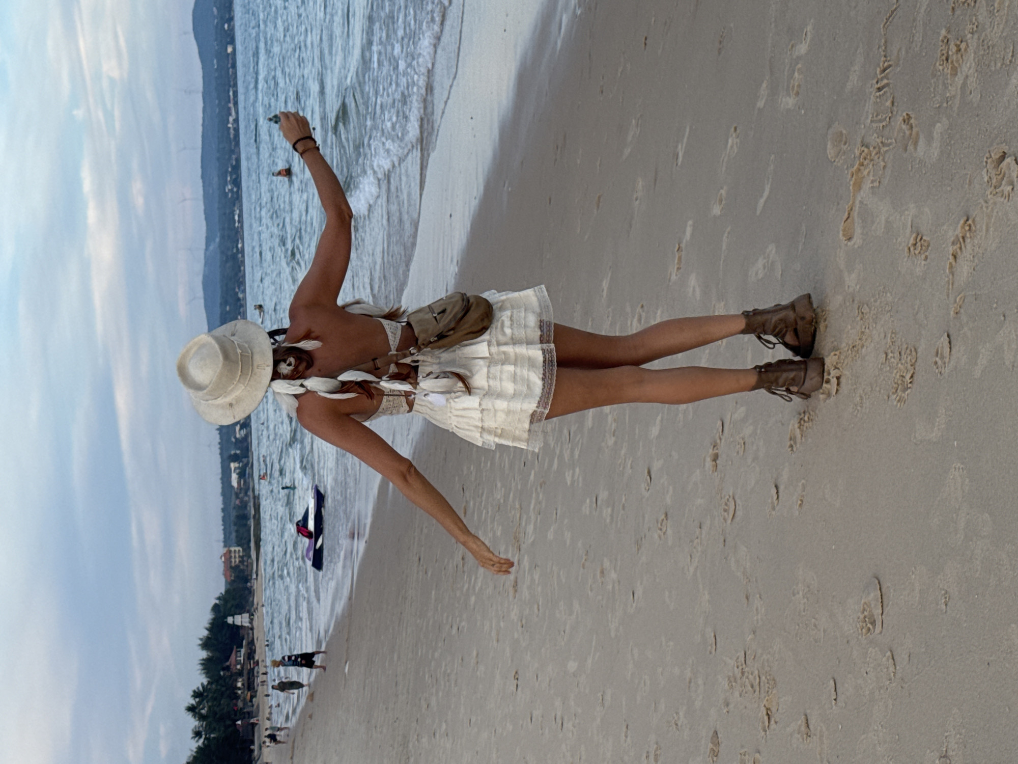 An individual, viewed from behind, stands on a sandy beach with their arms outstretched, facing the ocean. They wear a wide-brimmed hat, a white top with intricate detailing, and a short, tiered white skirt. A small, tan shoulder bag is slung across their back. Their legs are adorned with brown, open-toed lace-up boots. The water exhibits gentle waves, and a dark-colored jet ski is partially visible in the surf. In the distance, a shoreline features trees, buildings, and what appear to be wind turbines on a hill. The sky is overcast with a soft, diffused light. The sand is marked with numerous footprints and indentations. The scene is located on a beach in Phu Thuy Ward, Vietnam.