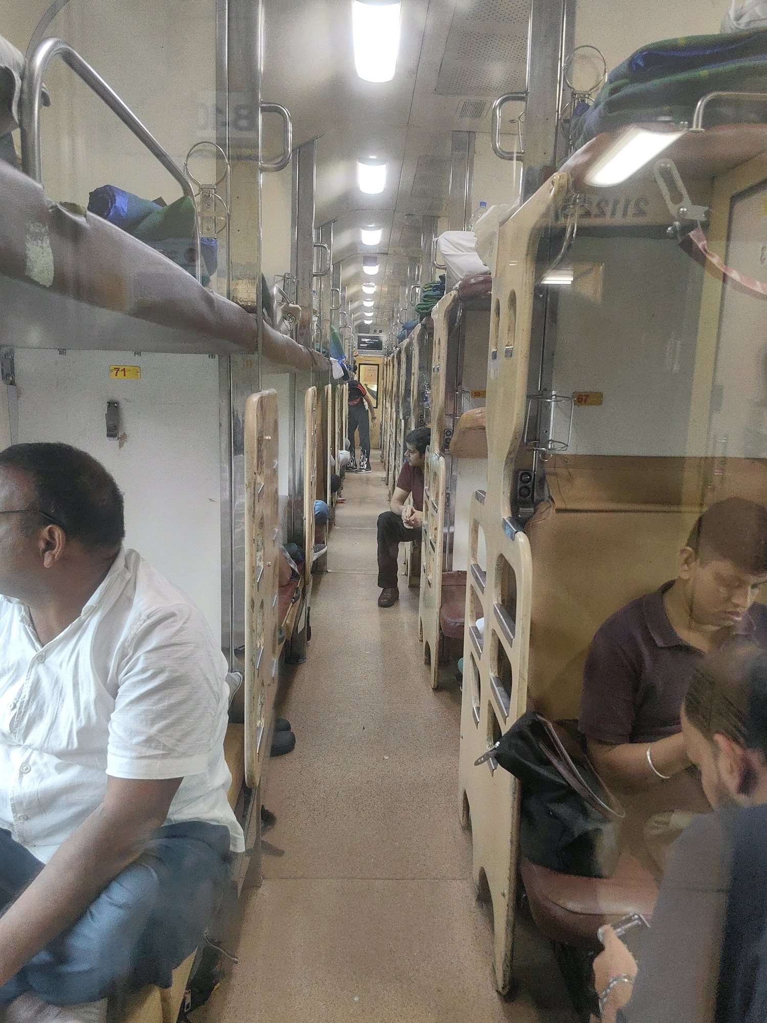 The image depicts the interior of a train carriage, possibly in Kelva, India, featuring multiple berths. In the foreground on the left, a man wearing glasses and a white short-sleeved shirt is seated. The aisle of the train is visible, lined with beige-colored berths, some occupied by individuals. Further down the aisle, another person is seen seated on a berth, looking down. The berths are stacked, indicating a sleeper class carriage. Lighting fixtures are visible along the ceiling of the carriage. The overall environment suggests a typical train journey within India.