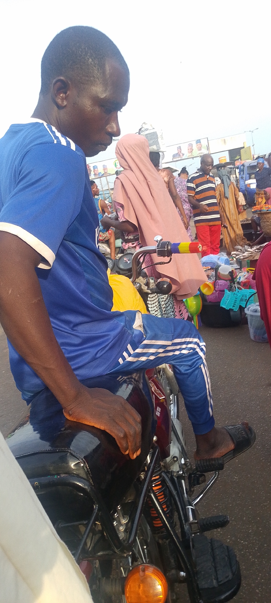 A man is depicted sitting on a motorcycle in what appears to be a bustling outdoor market in Lokoja, Nigeria. The man, with dark skin, is seen from his right side, looking downwards with a contemplative expression. He wears a blue short-sleeved t-shirt with white stripes on the sleeves and matching blue track pants with three white stripes along the side. His right hand rests on the motorcycle's black fuel tank, and he wears black slide sandals on his feet, one of which shows partially visible text, possibly a brand name like "BOSS."
The motorcycle features a black tank, and parts of its frame, suspension with orange springs, and an orange front indicator light are visible. In the background, the scene is vibrant with activity. A person in a light pink hijab or head covering is prominently positioned behind the man, possibly carrying items on their head. Further back, numerous individuals are present, including a man in an orange and brown striped shirt and red pants. Market stalls are visible with various goods, including colorful plastic items and textiles, arranged on the ground. The lighting suggests it is daytime, possibly late morning or afternoon, with a bright, hazy sky. In the far distance, several large political posters featuring multiple faces and unreadable text/logos are visible, indicating the urban or semi-urban setting. The overall atmosphere is that of a lively street or market environment.
