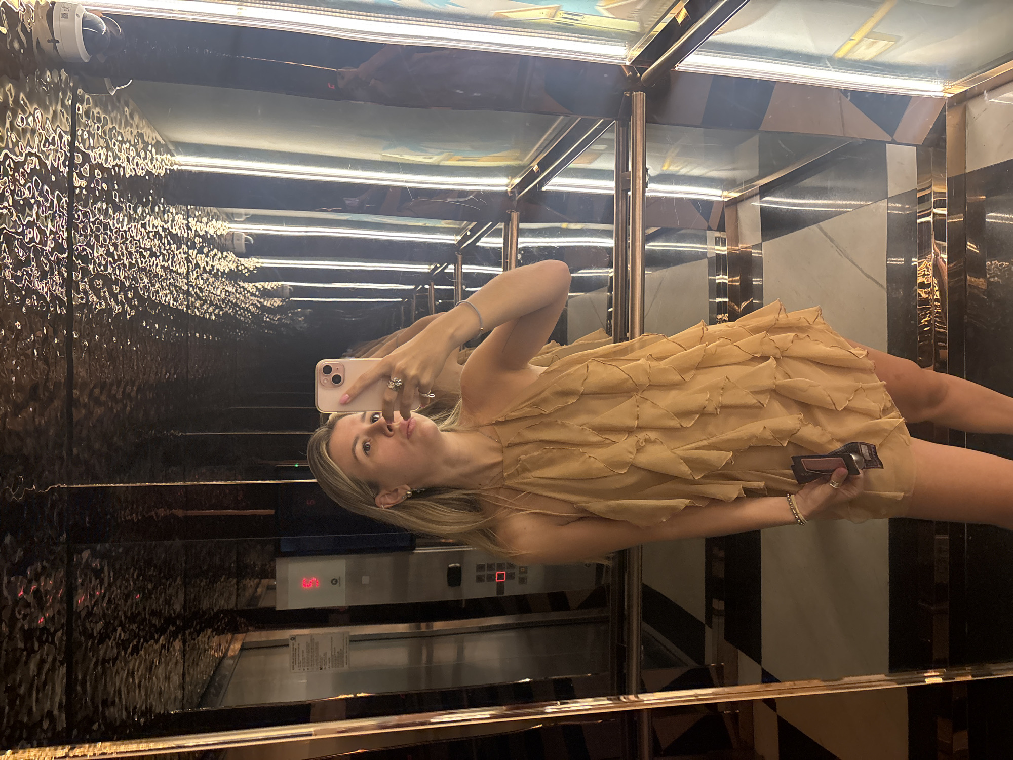 A young woman with blonde hair is taking a selfie in an elevator. She is wearing a tan, ruffled halter dress and several rings and bracelets. She is holding a cell phone and what appears to be a wallet in her right hand. The elevator has mirrored walls and a checkered floor. There are lights on the side walls of the elevator. The woman is looking at her phone with her lips puckered.