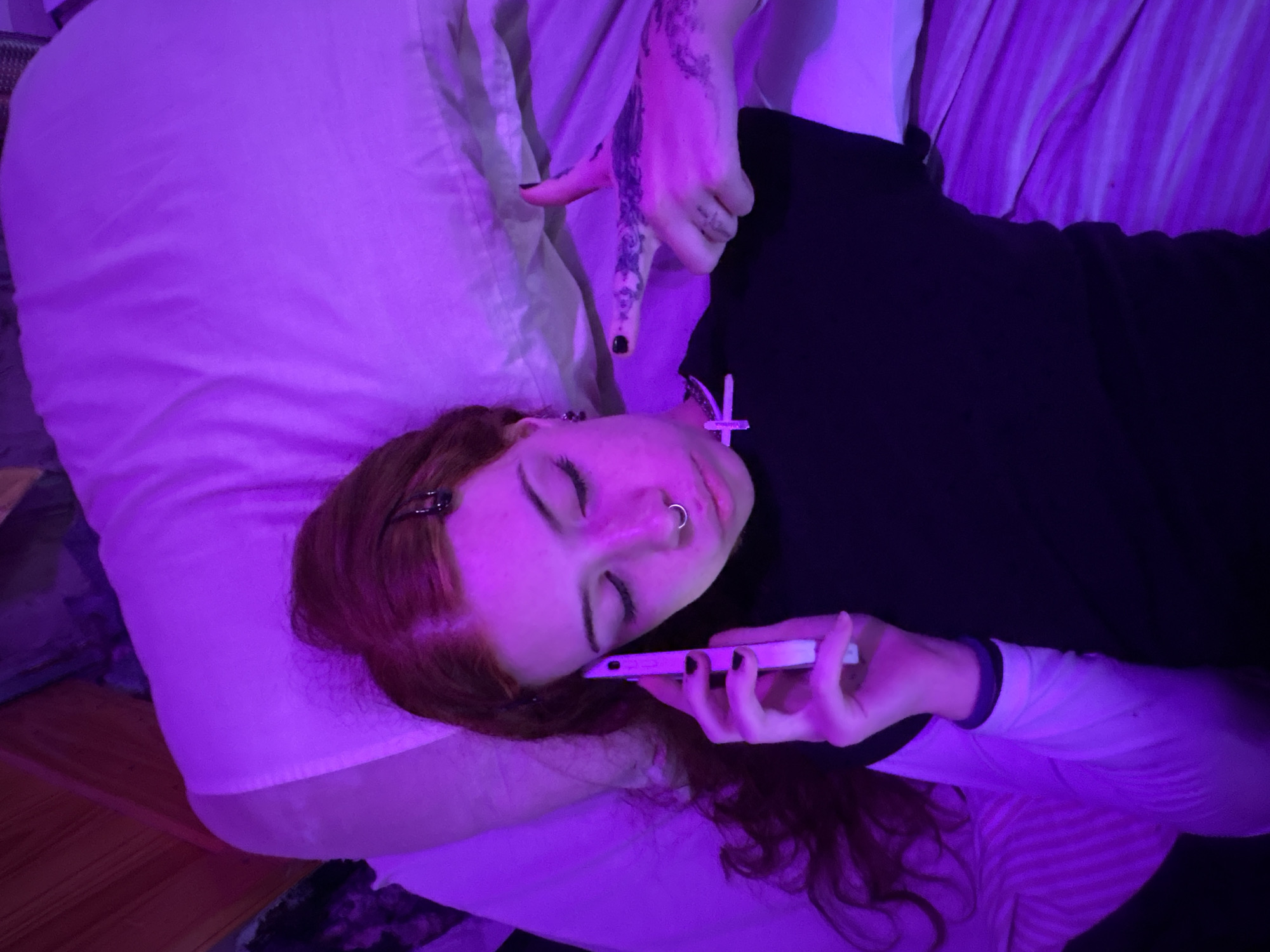 A person with red hair is lying on a bed, holding a smartphone to their face. They are wearing a black shirt and a white undershirt. Their eyes are closed. They have a nose ring and a small hair clip in their hair. Their right hand is tattooed. The lighting in the room is purple. The location is not discernible from the image.