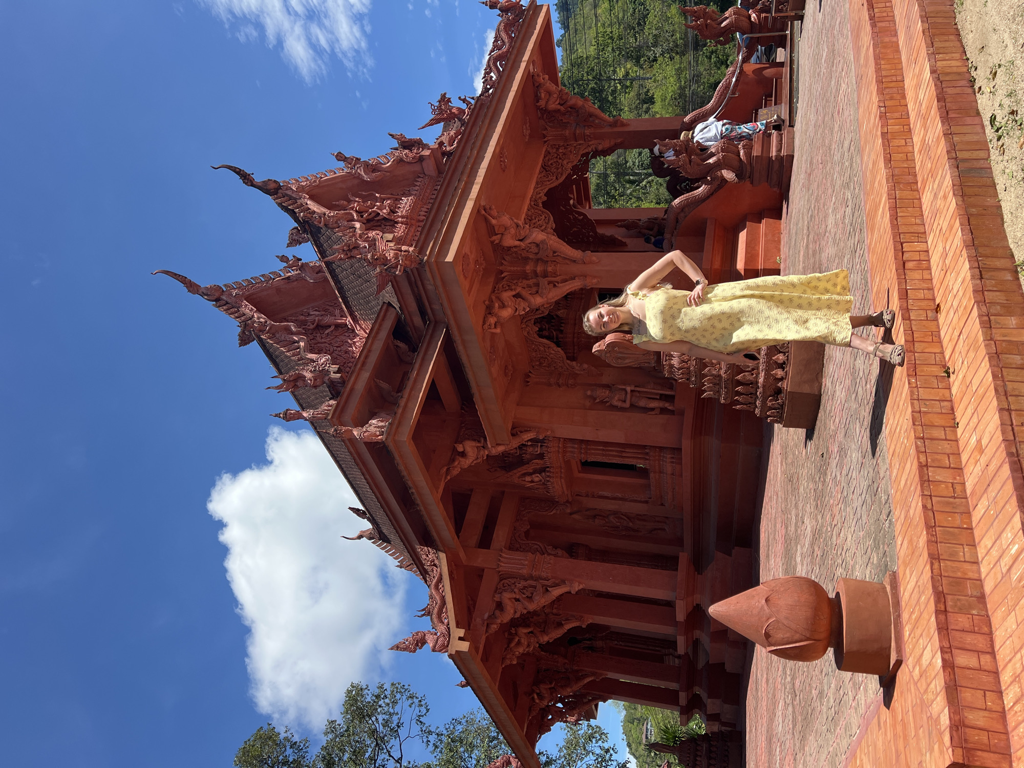 A full-length image shows a blonde woman wearing a floral yellow dress standing in front of Wat Sila Ngu (Red Temple) in Baan Hua Thanon, Thailand. She is standing to the right of the frame, with her left arm on her waist and her right arm down, and smiling at the camera. The temple is highly ornate with red-painted carvings and is made of brick. A blue sky with white clouds can be seen in the top left of the image.