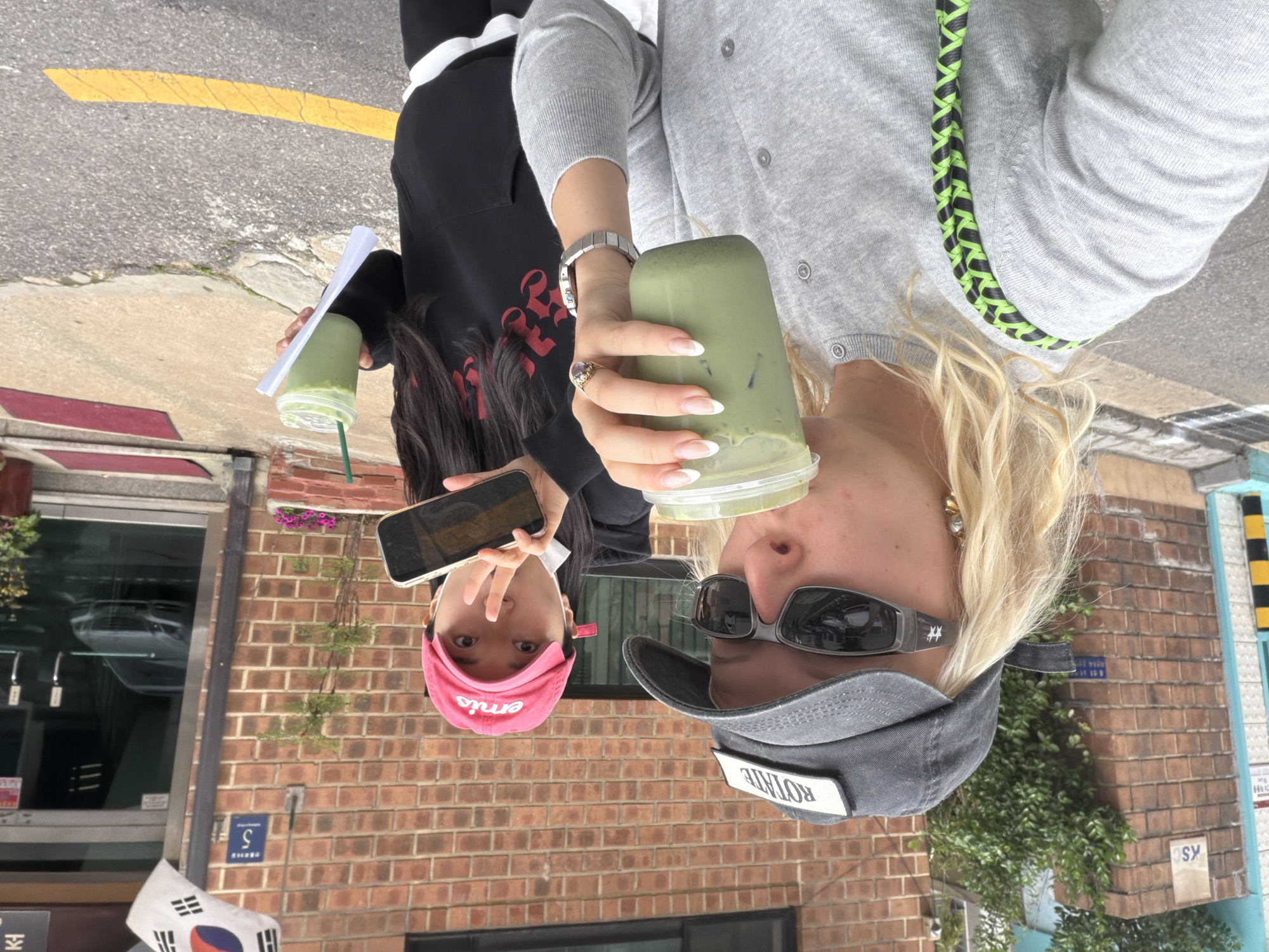 Two women are standing on a city street, each holding a green-colored drink in a plastic cup. The woman on the right is wearing a gray cardigan, sunglasses, and a gray cap with the word "ROTATE" on it. Her hair is blonde. She is holding the cup with her right hand, displaying a wristwatch and a ring. The woman on the left is wearing a black hoodie with red details and a pink cap with the word "Emis" on it. She is holding a phone in her left hand and making a peace sign with her right hand.
Behind them is a brick building. A South Korean flag is to the left, near the bottom of the image. A small street sign with the number "5" is visible on the brick wall. Another sign, "SY," is on a pillar. Foliage grows along the building's exterior.
The setting is urban. A yellow line is painted on the asphalt road, indicating the presence of traffic. The location is Seoul, South Korea.