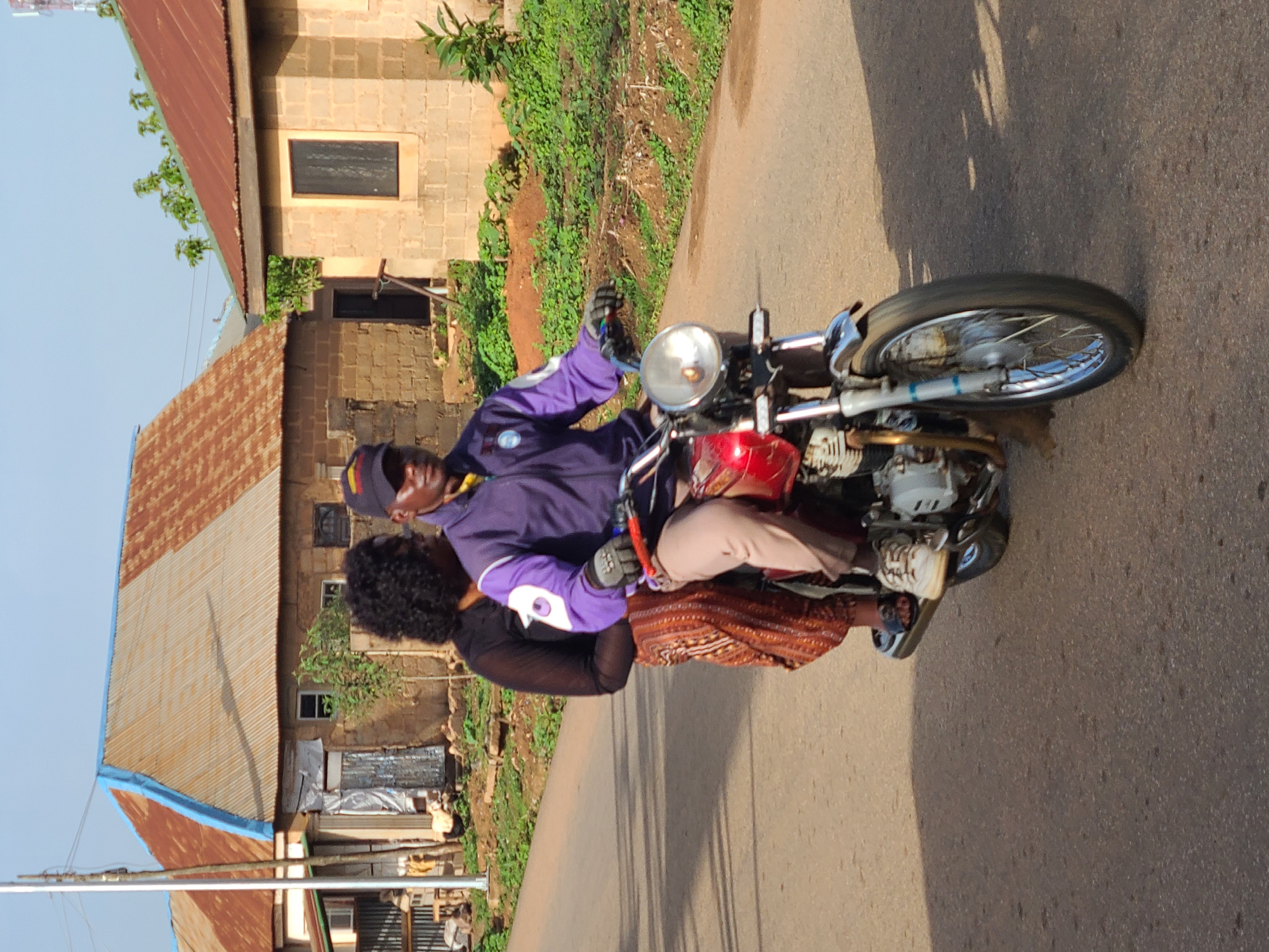 On a paved road in an urban area identified as Unknown, Unknown, a man and a woman are riding a red motorcycle. The man, wearing a purple jacket and a cap, is operating the motorcycle, while the woman is seated behind him. Adjacent to the road, there are buildings with corrugated metal roofs, greenery, and soil. The time of day appears to be daytime, with clear lighting.