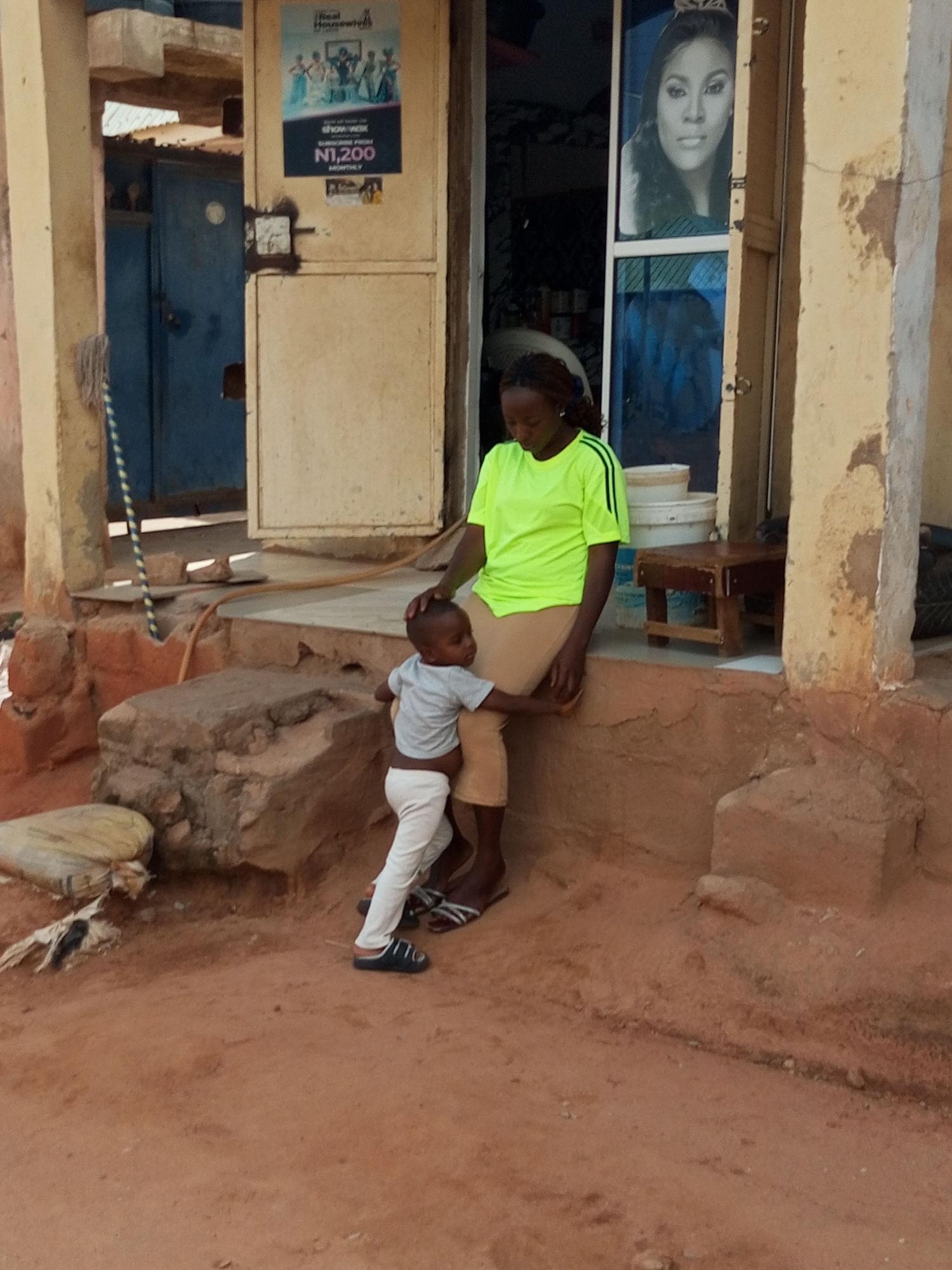 In Gombe, Nigeria, a woman in a bright neon green shirt and beige pants sits on a raised step outside a building. She is holding a young child who is wearing a light gray t-shirt and white pants. The child stands beside her, facing away from the camera, with one hand resting on the woman's knee. The woman's other hand is resting on the child's back.
Behind them, a door is visible with a poster featuring a woman's face. Another poster on the door advertises "Real Housewives" with a price of "N1,200 MONTHLY." To the left of the door is a blue, metal-clad structure with a mop leaning against it. The building appears to be constructed with weathered materials, with portions of the wall showing exposed brick and rough texture. A small wooden stool is placed near the door. The ground in front of the building is sandy and reddish-brown. The overall scene suggests an outdoor setting in a possibly rural or less developed urban area. The lighting indicates it is daytime.