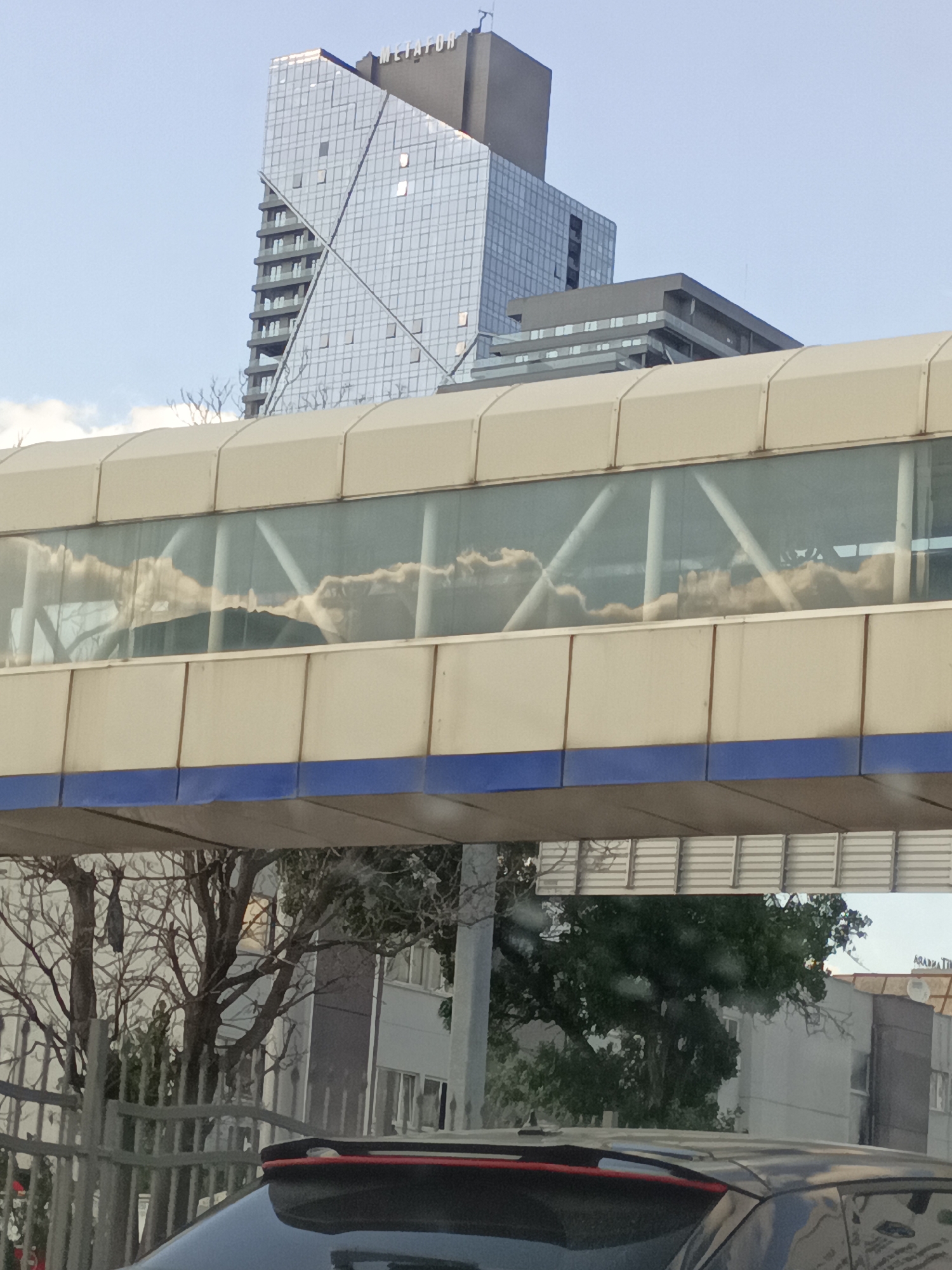 In Altındağ, Turkey, a modern pedestrian overpass with a glass enclosure spans across a street. The glass reflects the sky and clouds, creating a distorted view of the environment. In the background, a contemporary skyscraper with a unique angular design dominates the skyline, its facade covered in reflective glass panels. Below the overpass, a metal fence partially obstructs the view of trees and buildings. A dark-colored car is visible in the foreground, with its roof and spoiler partially in frame. The scene captures an urban environment characterized by modern architecture and reflections.
