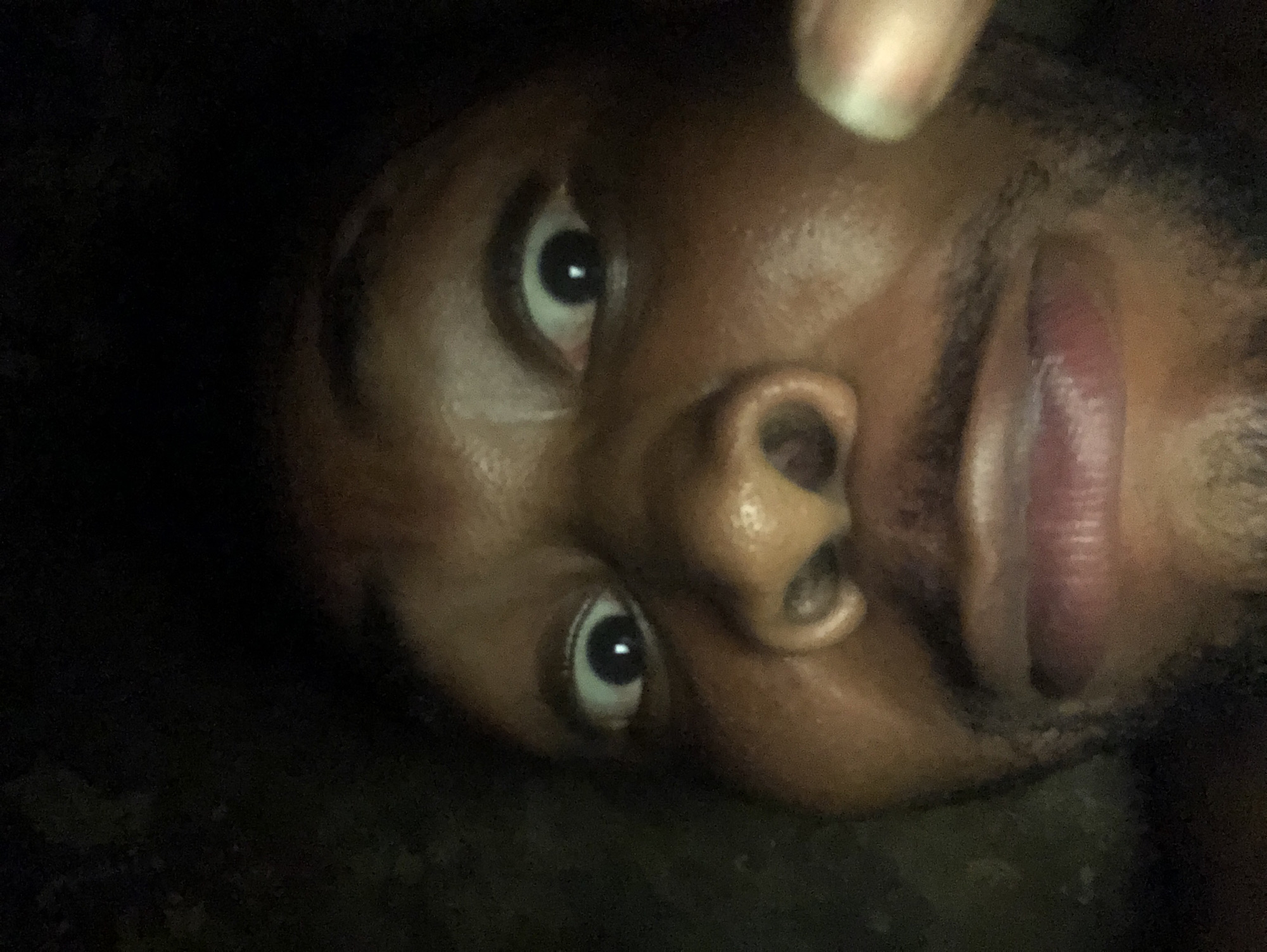 A close-up image of a person's face, likely male with dark skin, is seen against a dark background. The face is slightly rotated. The subject has dark eyes and a prominent nose. Part of a finger is visible at the top. The location is Unknown, Unknown.
