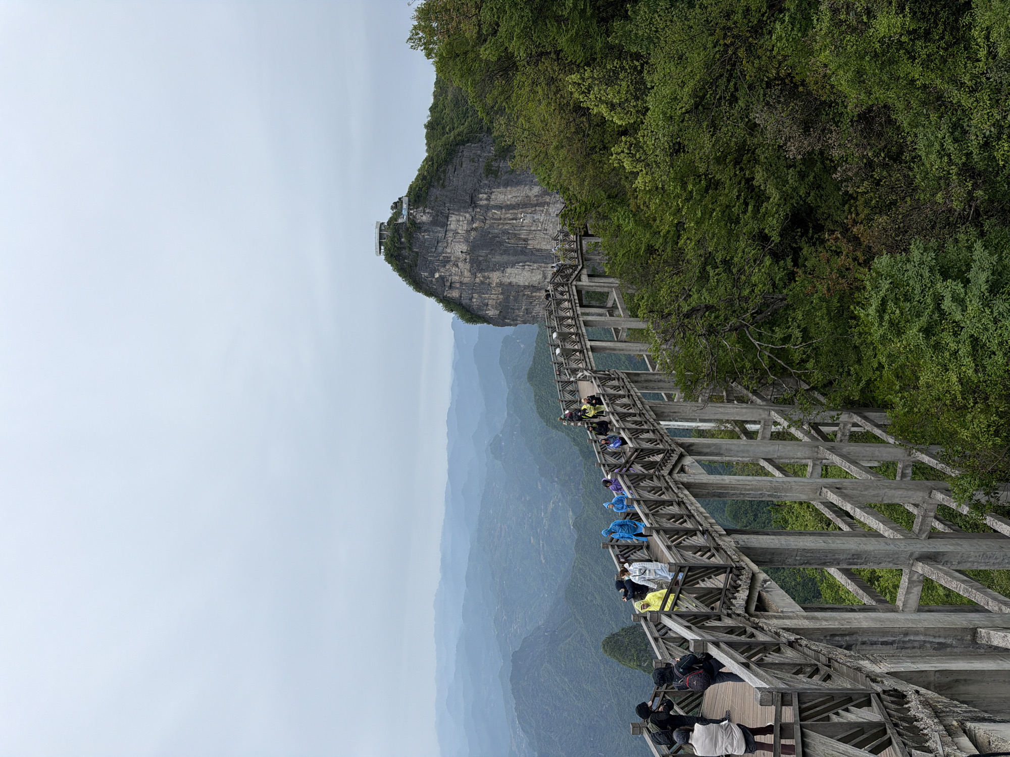 The image shows a high-altitude walkway at Tianmen Mountain in Zhangjiajie, China. The walkway, constructed of light-colored wooden beams, is built along the side of a cliff face. People wearing various colored raincoats and jackets are visible along the walkway. In the background, layered mountain ranges are visible through a hazy atmosphere. The top of the cliff has some structures, possibly a viewing platform. Trees and vegetation cling to the cliff edge. The sky is overcast.