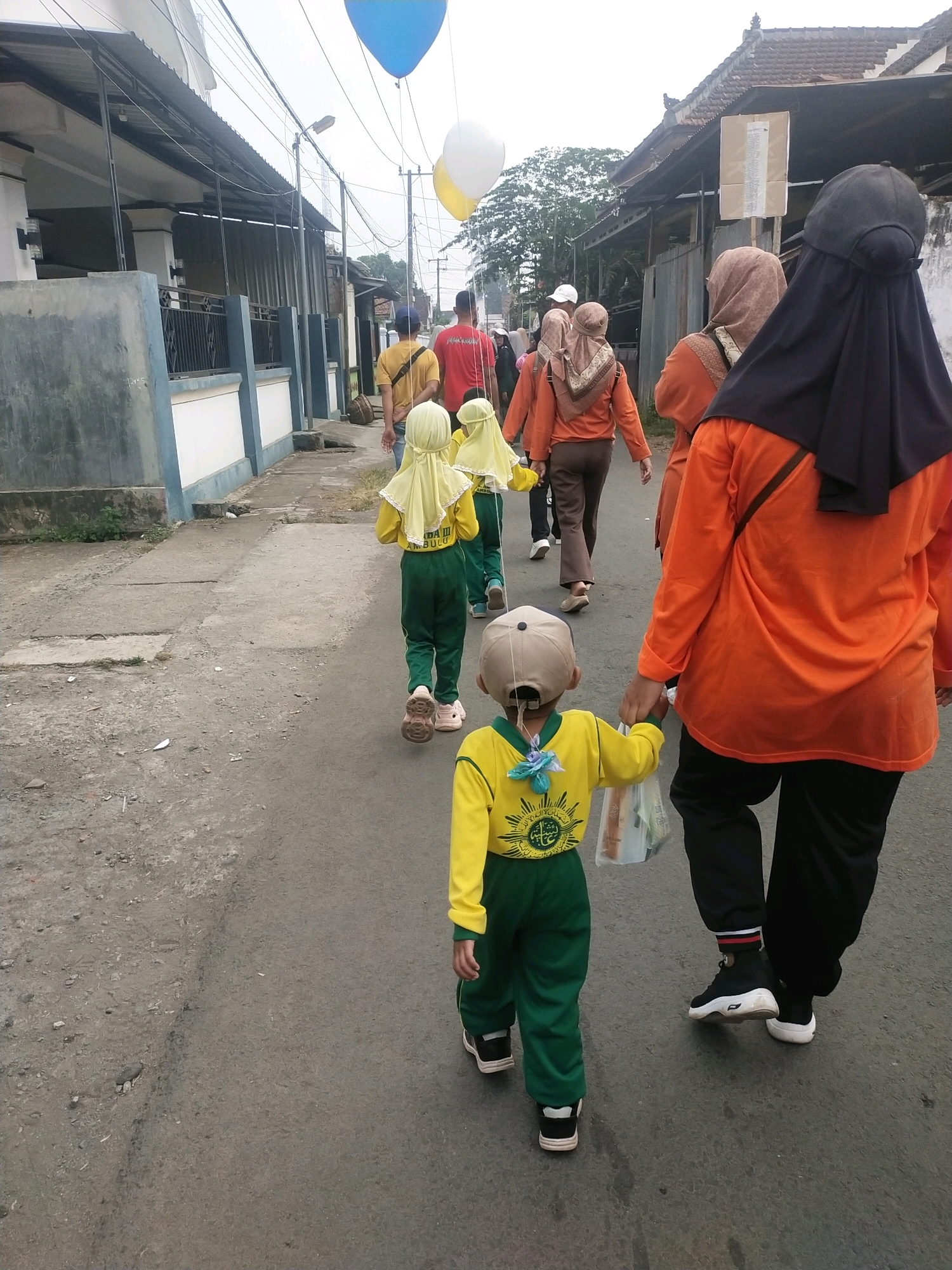 In Ambulu, Indonesia, a group of people, predominantly women and children, walk along a paved street lined with residential buildings. The scene is dominated by figures from behind, indicating a procession or walk. A young child in a yellow and green uniform, wearing a baseball cap, leads the way, holding a plastic bag. Behind this child, other children in similar yellow uniforms and headscarves are visible, followed by adults. Several women are wearing headscarves and loose-fitting clothing in orange and brown. Two large balloons, one blue and one yellow and white, are suspended in the air, suggesting a celebratory or event-related occasion. The street is made of asphalt and shows signs of wear, with some debris present. The buildings on either side of the street appear to be simple, single-story structures with tiled roofs. Power lines and utility poles are visible overhead. The overall impression is of a community gathering in a semi-urban environment.