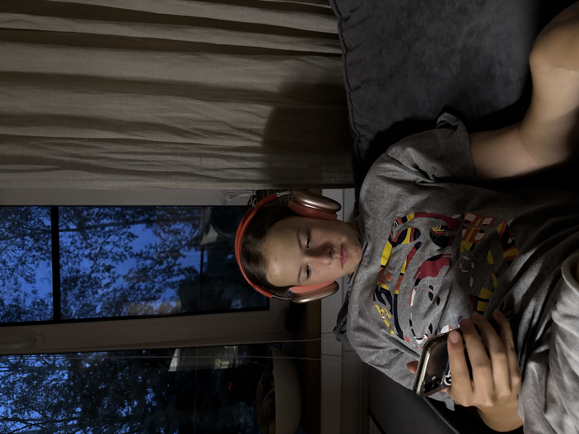 An indoor scene captures a young person wearing orange over-ear headphones, engrossed in their smartphone. They are seated on a dark gray couch, their body partially obscured by a gray blanket. The person is wearing a gray t-shirt adorned with a colorful abstract pattern. In the background, a window reveals a twilight sky with trees silhouetted against it. To the left of the window, a white bowl filled with bananas sits on a surface. A light-colored curtain hangs to the right of the window, partially covering the view. The overall lighting suggests it is evening. The image was taken in Chaikovsky, Russia.