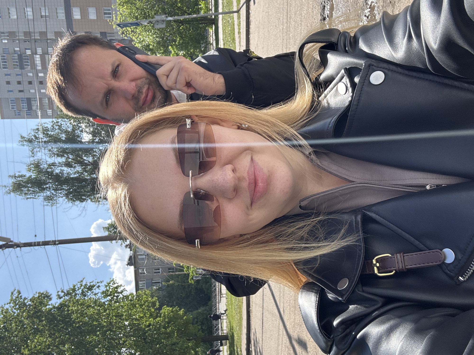 A woman with light blonde hair and sunglasses, wearing a black leather jacket, is taking a selfie with a man partially visible behind her. The man is on a phone call, holding the phone to his ear. The background shows a street scene with trees, buildings, and power lines. Based on the buildings and style, this is most likely in Russia. The specific location is Rusanovo in Russia.