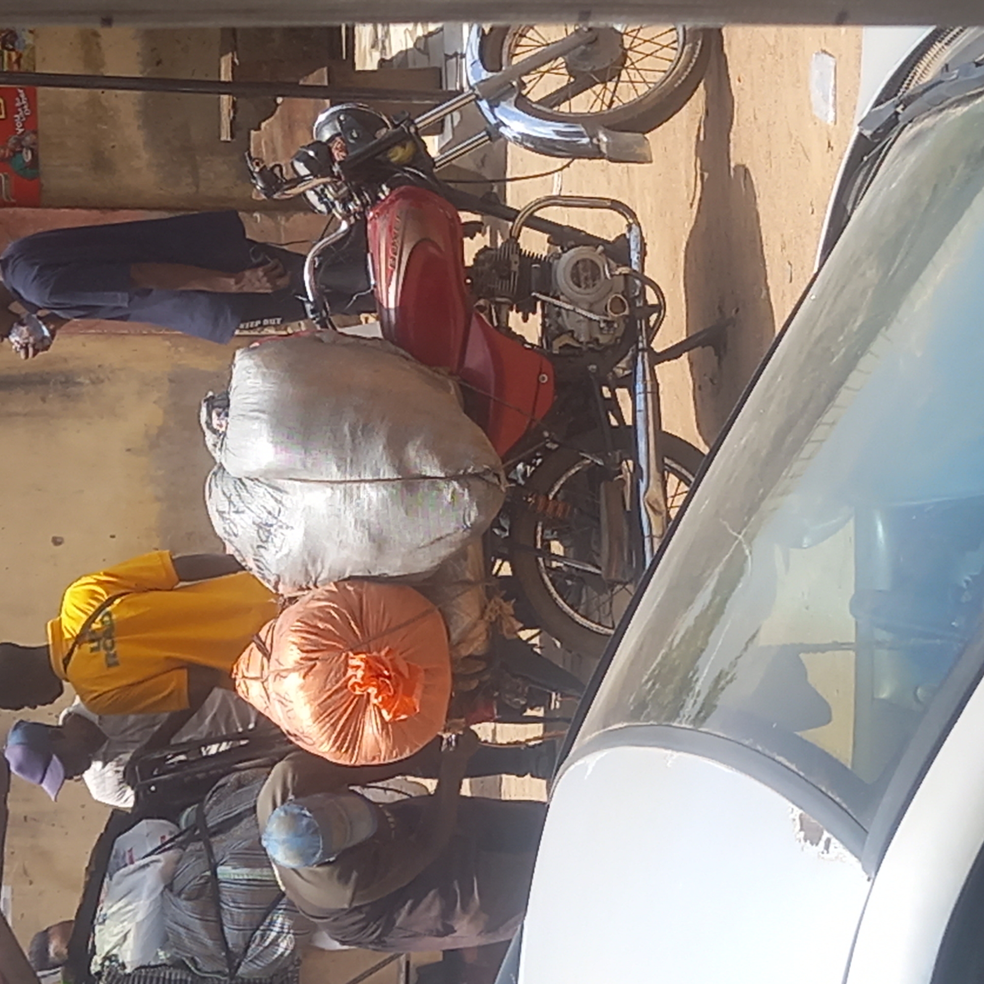 In Dadi Riba, Nigeria, a motorcycle is heavily laden with cargo including large bags. Several individuals are nearby, one wearing a yellow shirt. The image is taken from inside a vehicle, with the windshield visible. The scene suggests a busy urban environment.