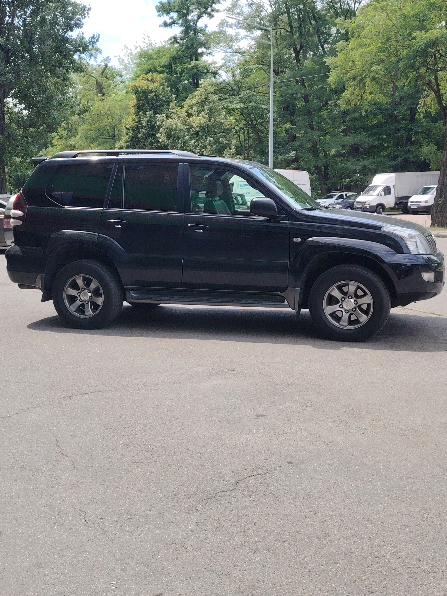 A black Toyota Land Cruiser Prado is parked on an asphalt road. The vehicle is viewed from the side, facing right. The car has a roof rack, tinted windows, and alloy wheels. In the background, there are trees, a street lamp, and other vehicles, including a white van and a white truck. The scene appears to be a street in Kyiv, Ukraine, given the context. The sky is partly cloudy and the lighting suggests it is daytime.